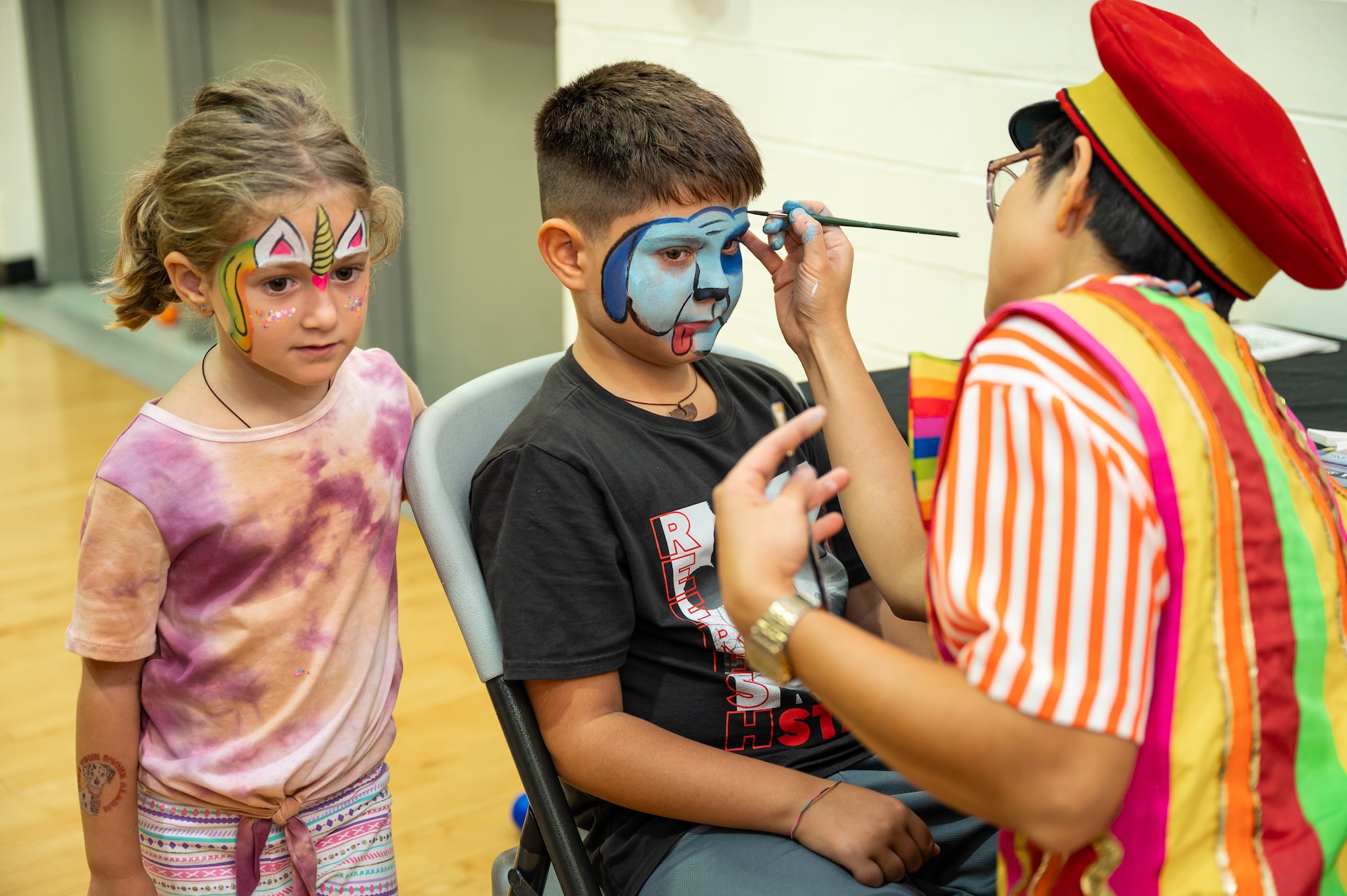 A child with unicorn face paint stands beside a seated boy getting a blue dog face painted by an artist in a red hat and colorful striped vest.