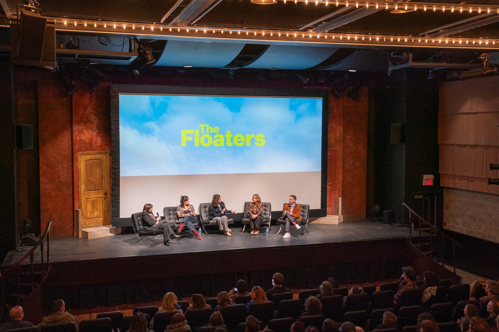Panel of five people seated on stage in front of a screen displaying 'The Floaters' with an audience watching.