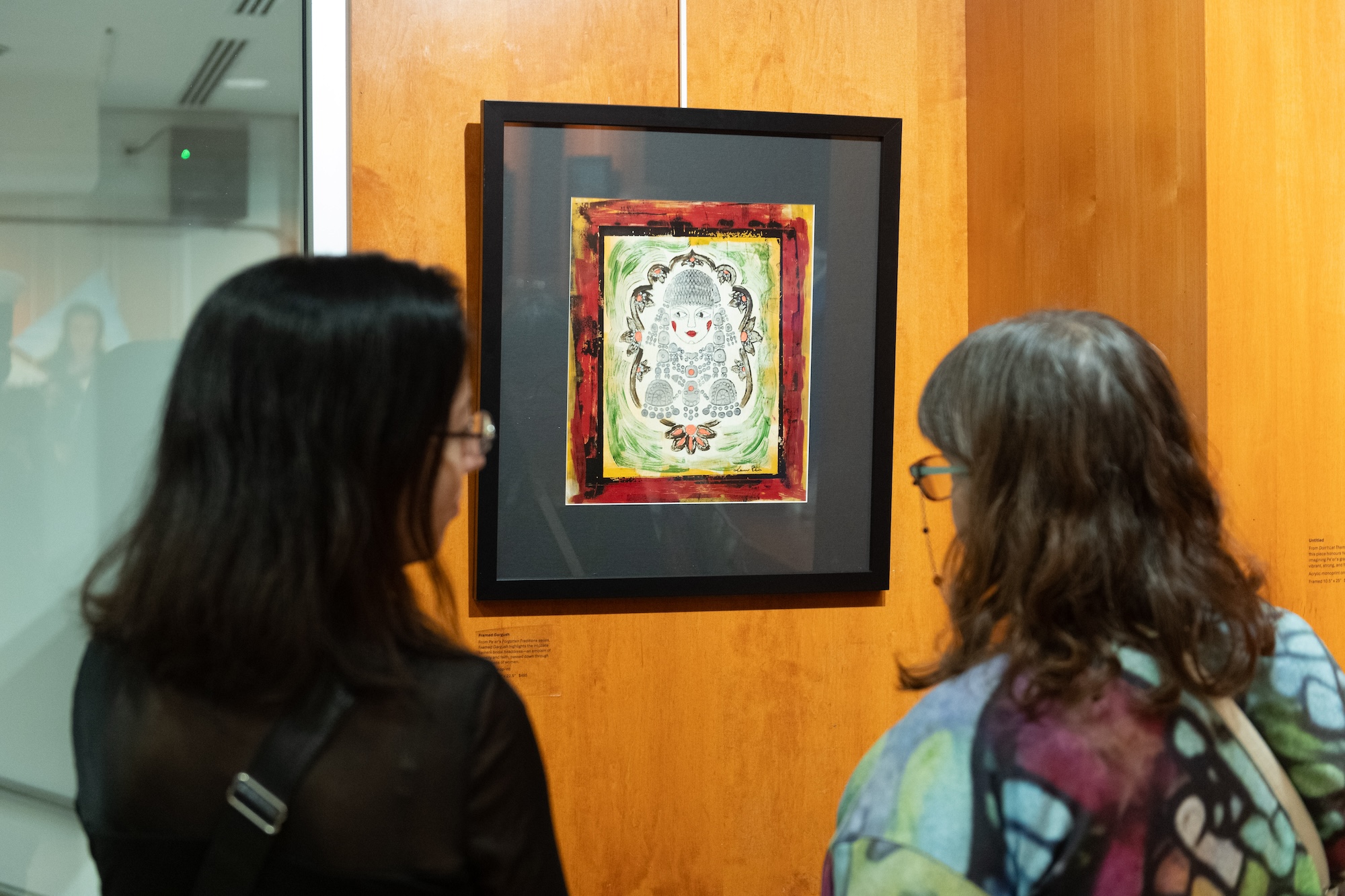 Two women with glasses viewing a colorful framed artwork featuring a stylized face on a wood-paneled gallery wall.