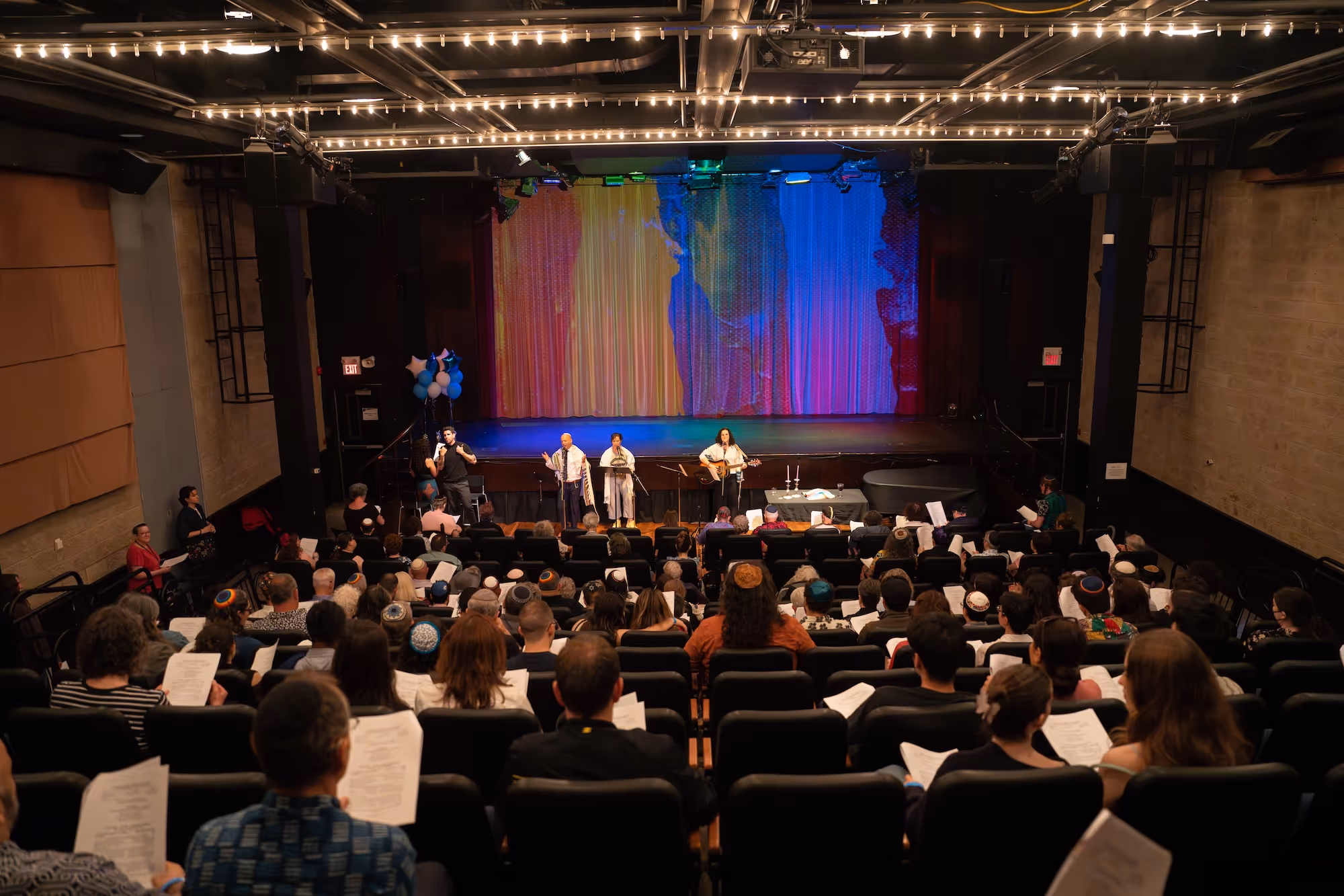 Audience seated in a theater watching four performers on stage with colorful curtains and string lights overhead.