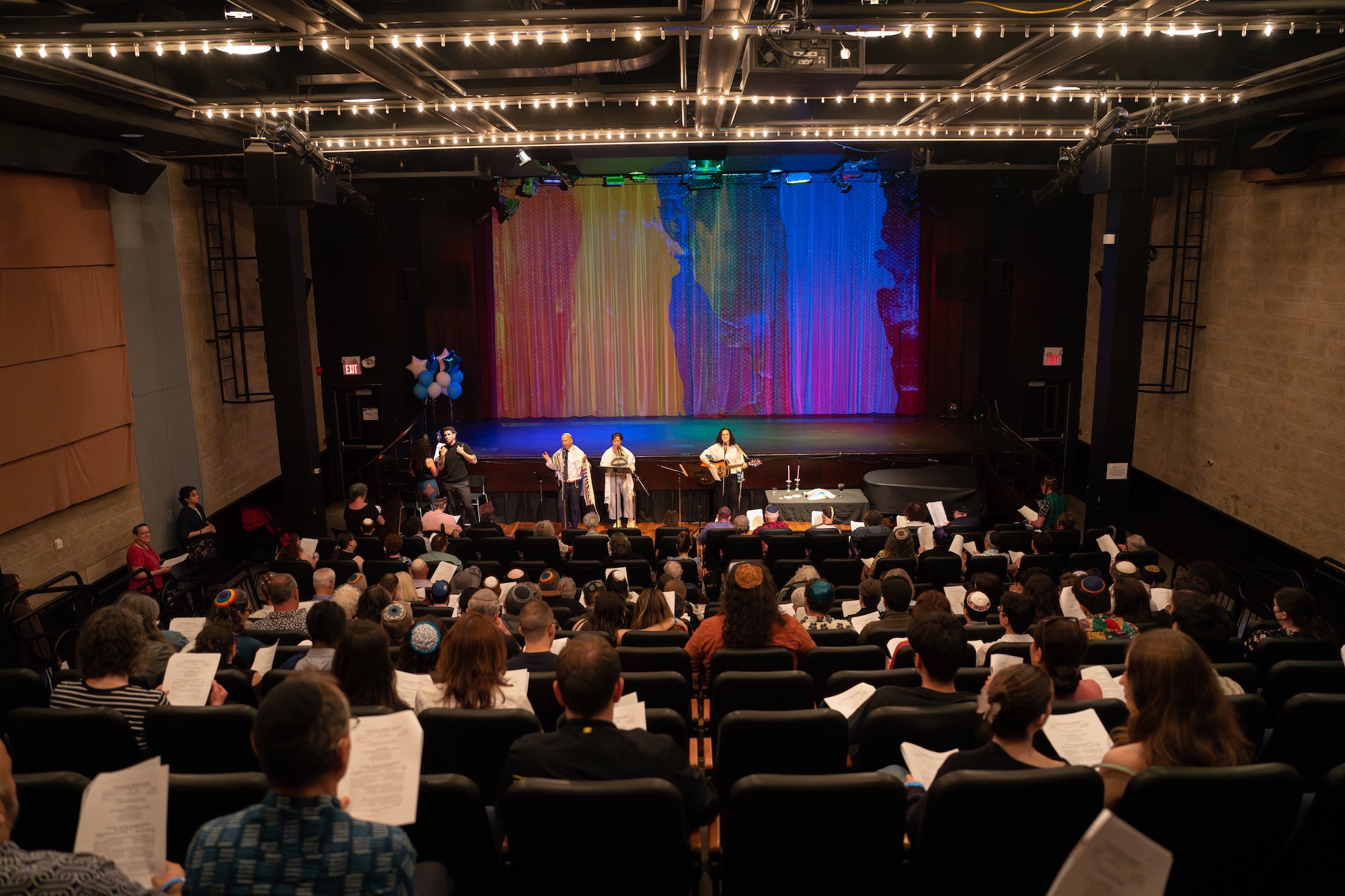 Audience seated in a theater watching four performers on stage with colorful curtains and string lights overhead.