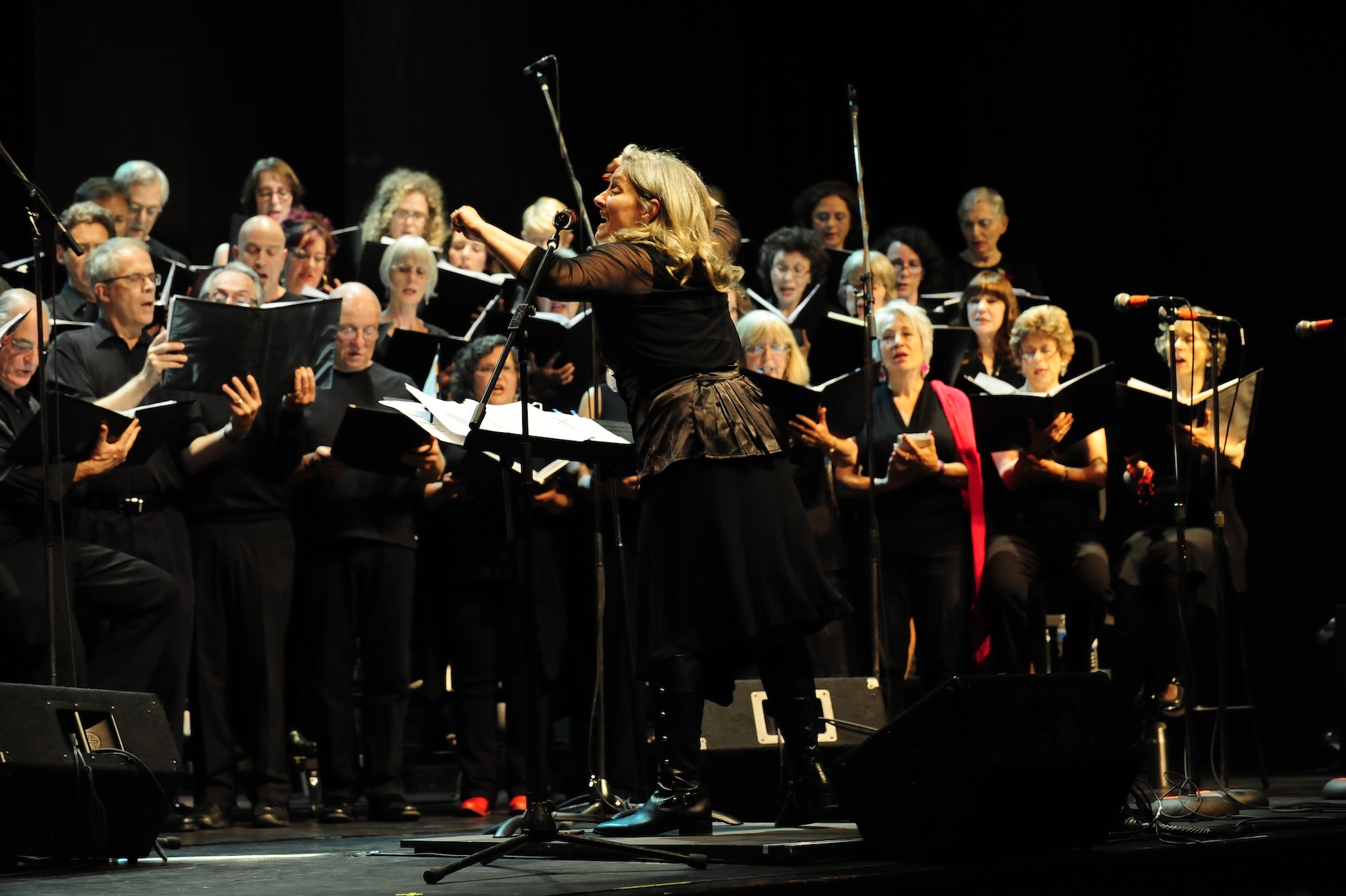 Female conductor leading a choir of adults singing on stage in a dark setting.