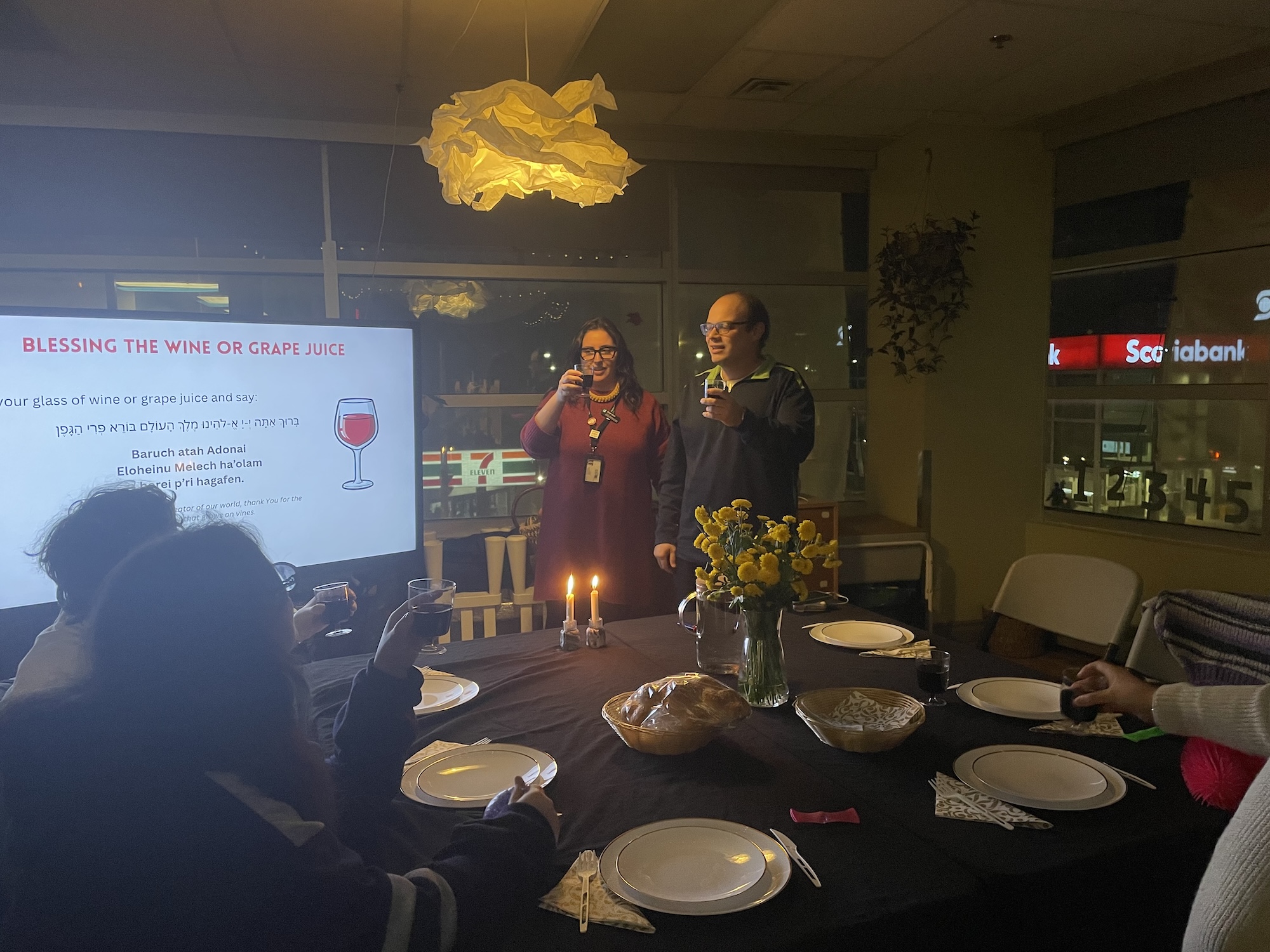 Group of people raising glasses to do the blessing over the wine before shabbat dinner.  A table set with plates, candles, flowers, and bread, with a screen showing the blessing text.