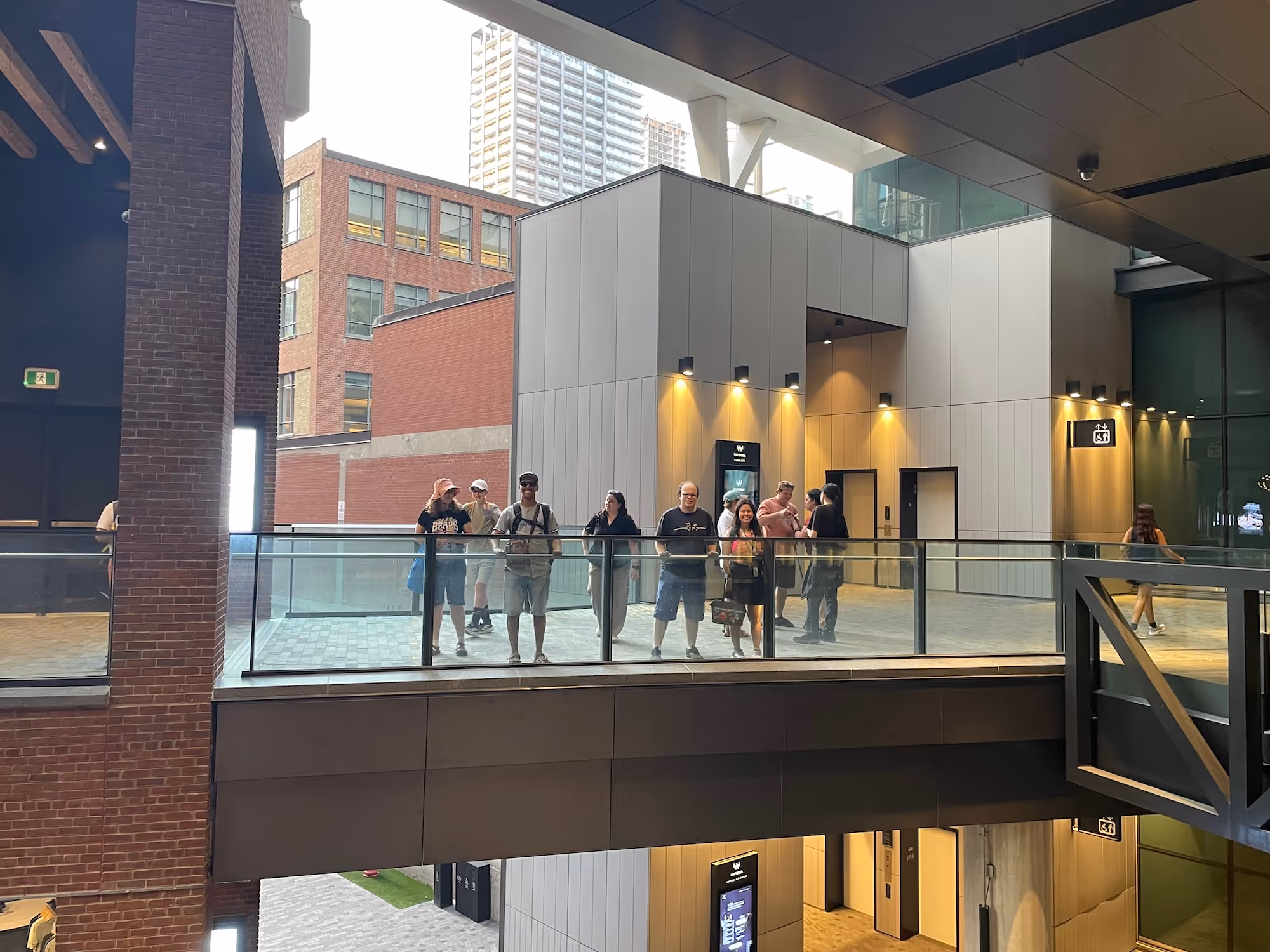 Group of people standing on a modern indoor balcony with glass railing inside a building, brick and concrete walls in background.