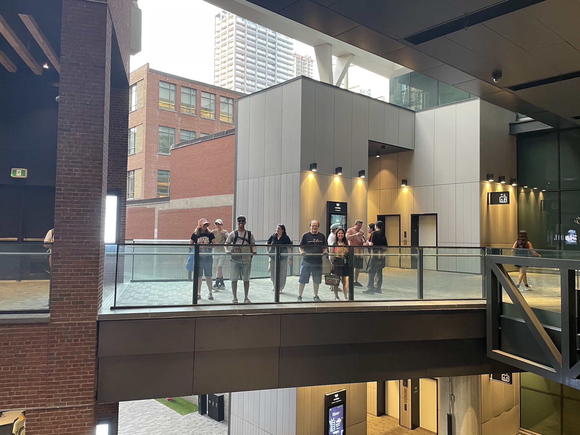 Group of people standing on a modern indoor balcony with glass railing inside a building, brick and concrete walls in background.