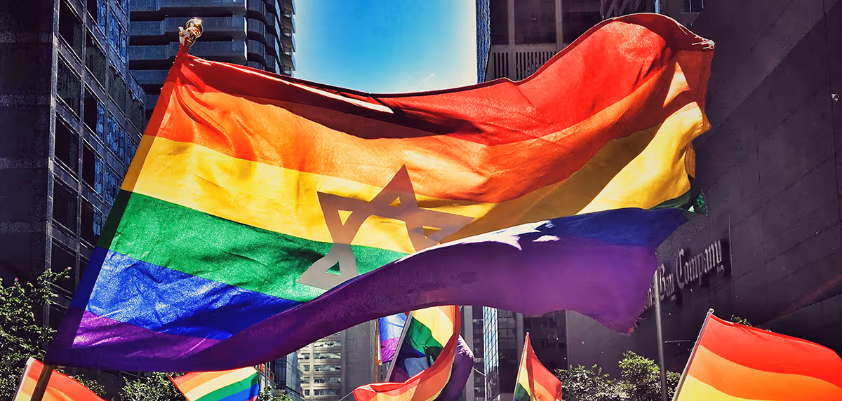 Large rainbow LGBTQ+ pride flag with a translucent Star of David waving above smaller rainbow flags in a cityscape with tall buildings.