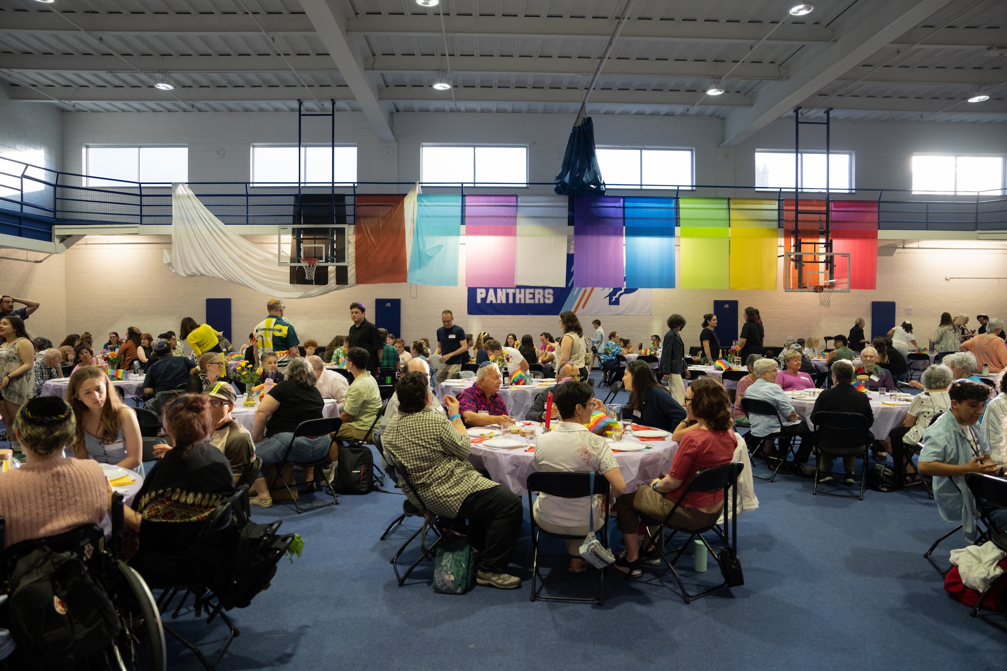 Large group of people seated at round tables inside a gymnasium decorated with colorful hanging flags and a 'Panthers' banner.