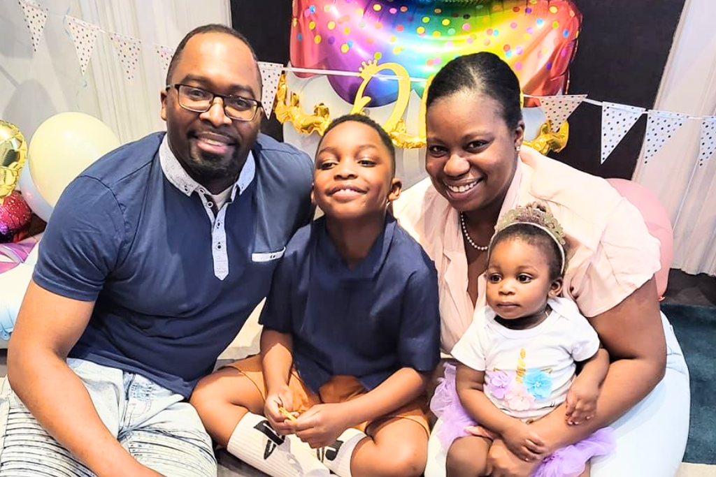 Smiling family of four sitting closely together at a birthday celebration with colorful balloons and decorations in the background.