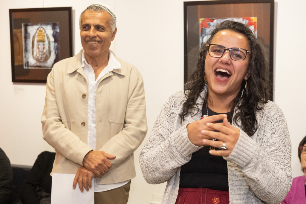 Two people standing and smiling, one speaking joyfully with hands clasped, in an indoor setting with framed artwork in the background.