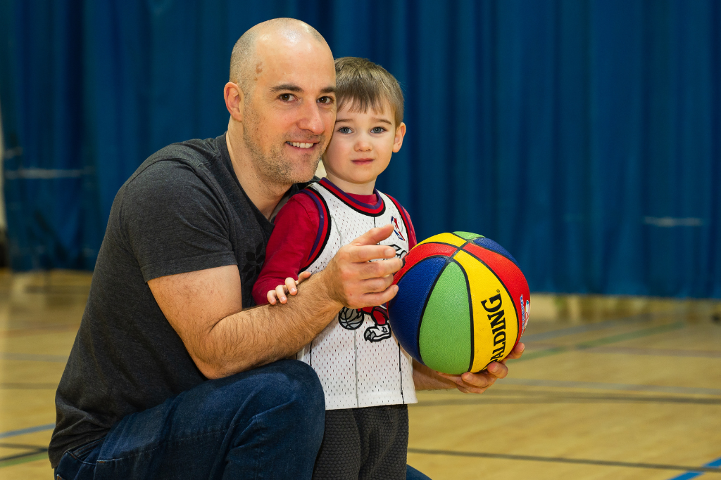 Adult man kneeling beside young boy holding a colorful basketball in a gymnasium.