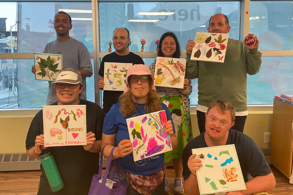A group of seven smiling adults holding up colorful artwork featuring leaves and painted designs in a bright indoor space.