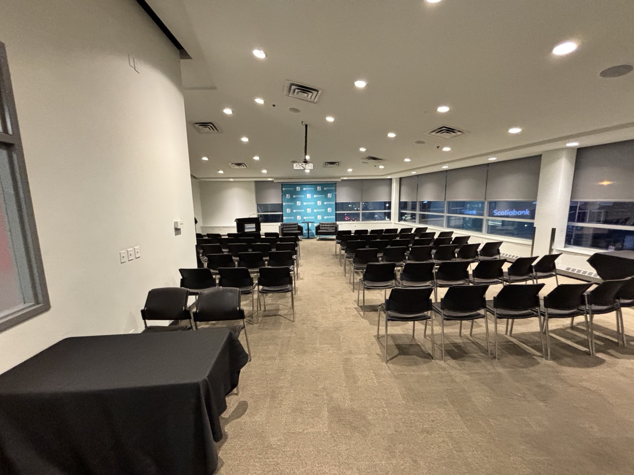 Large conference room with rows of black chairs facing a stage area with a blue backdrop and two armchairs, with windows on the right side.