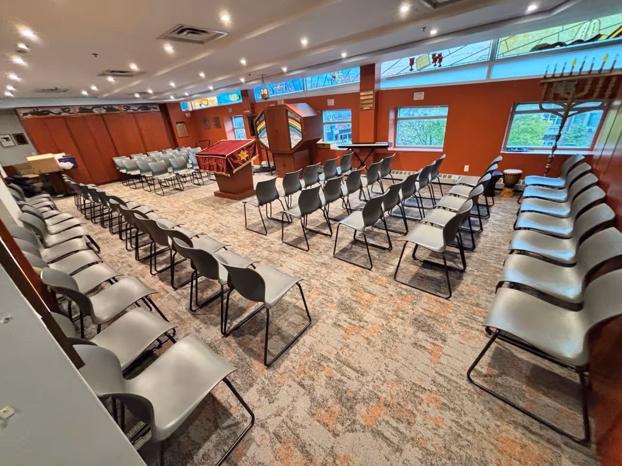 Spacious chapel with rows of gray chairs arranged facing a podium draped with a maroon cloth featuring a gold star, stained glass windows, and an orange accent wall.
