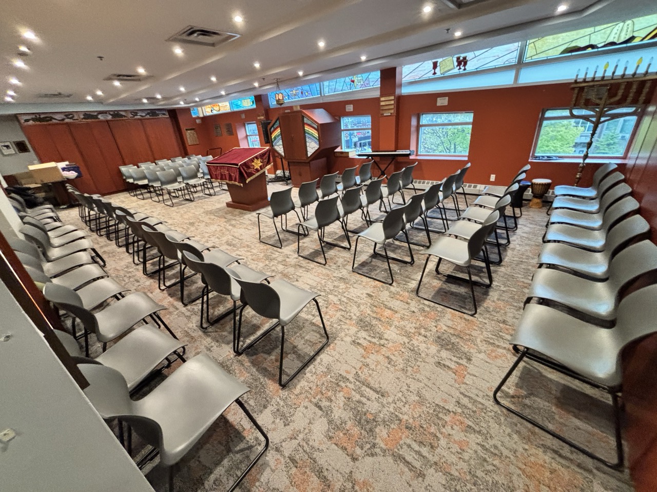 Spacious chapel with rows of gray chairs arranged facing a podium draped with a maroon cloth featuring a gold star, stained glass windows, and an orange accent wall.