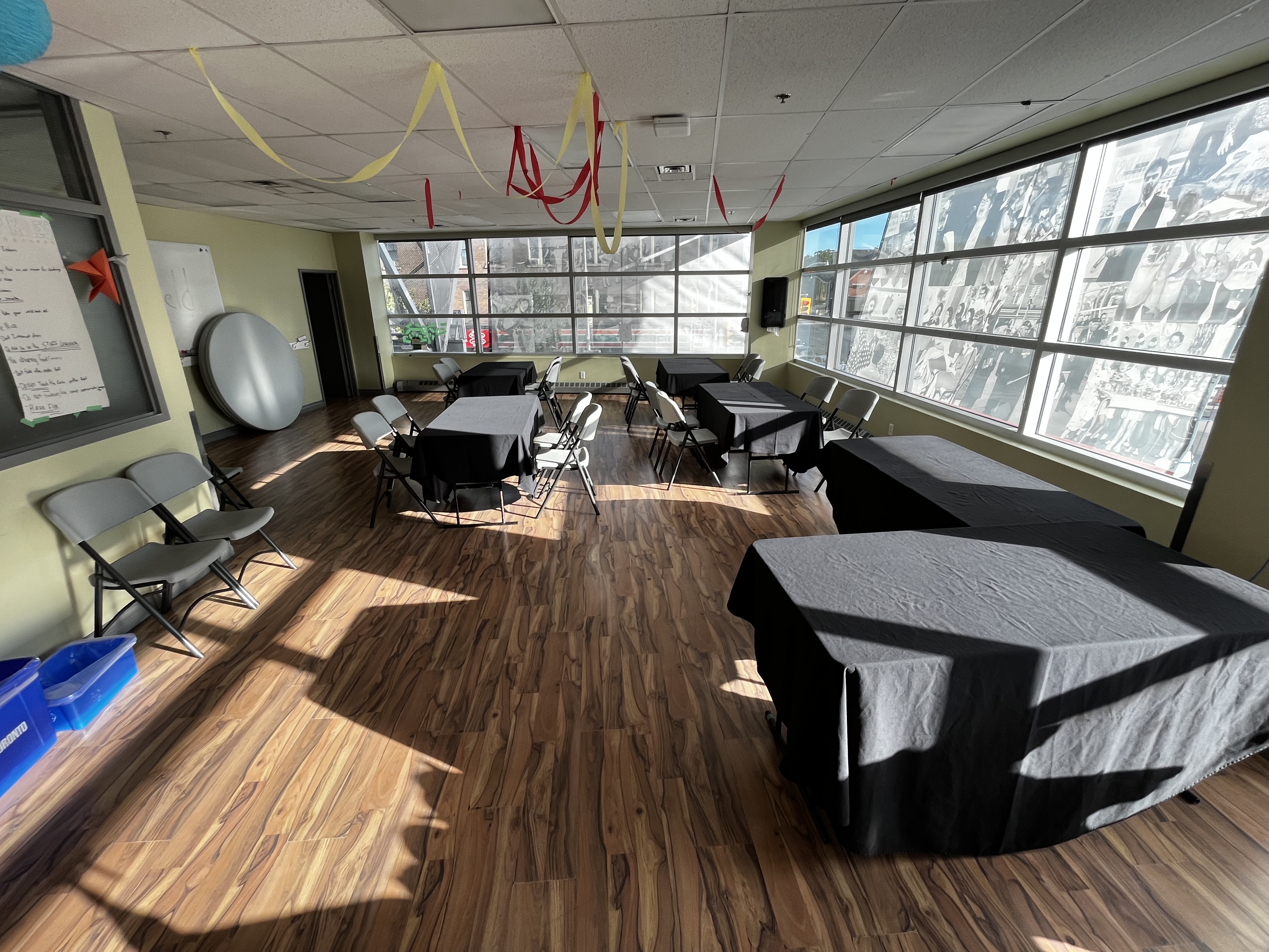 Sunlit room with multiple tables covered in black cloth and folding chairs arranged around them, with large windows displaying black-and-white historical photos.