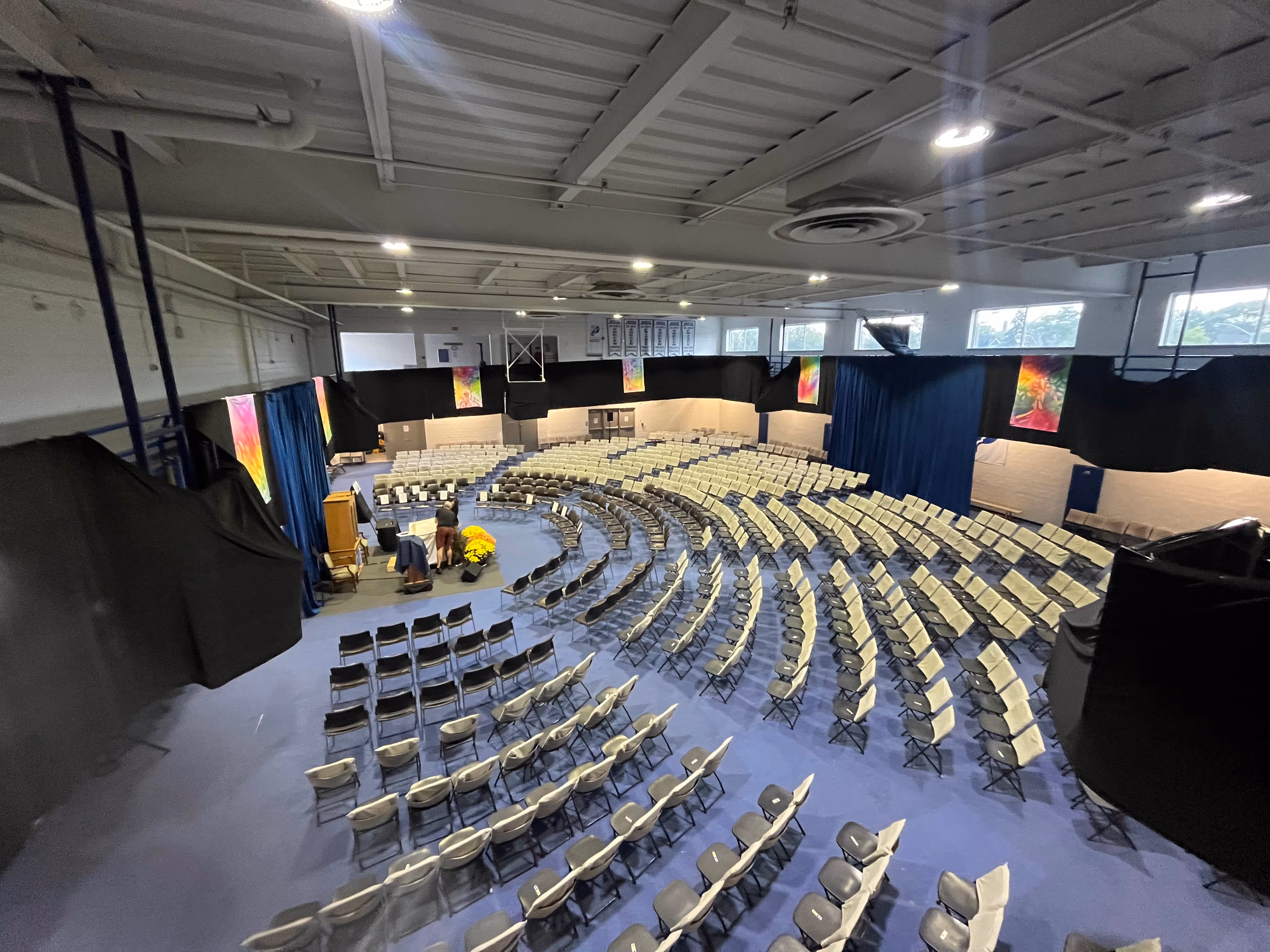 Large indoor auditorium with rows of folding chairs arranged in a semicircle facing a stage with a podium and yellow flowers.