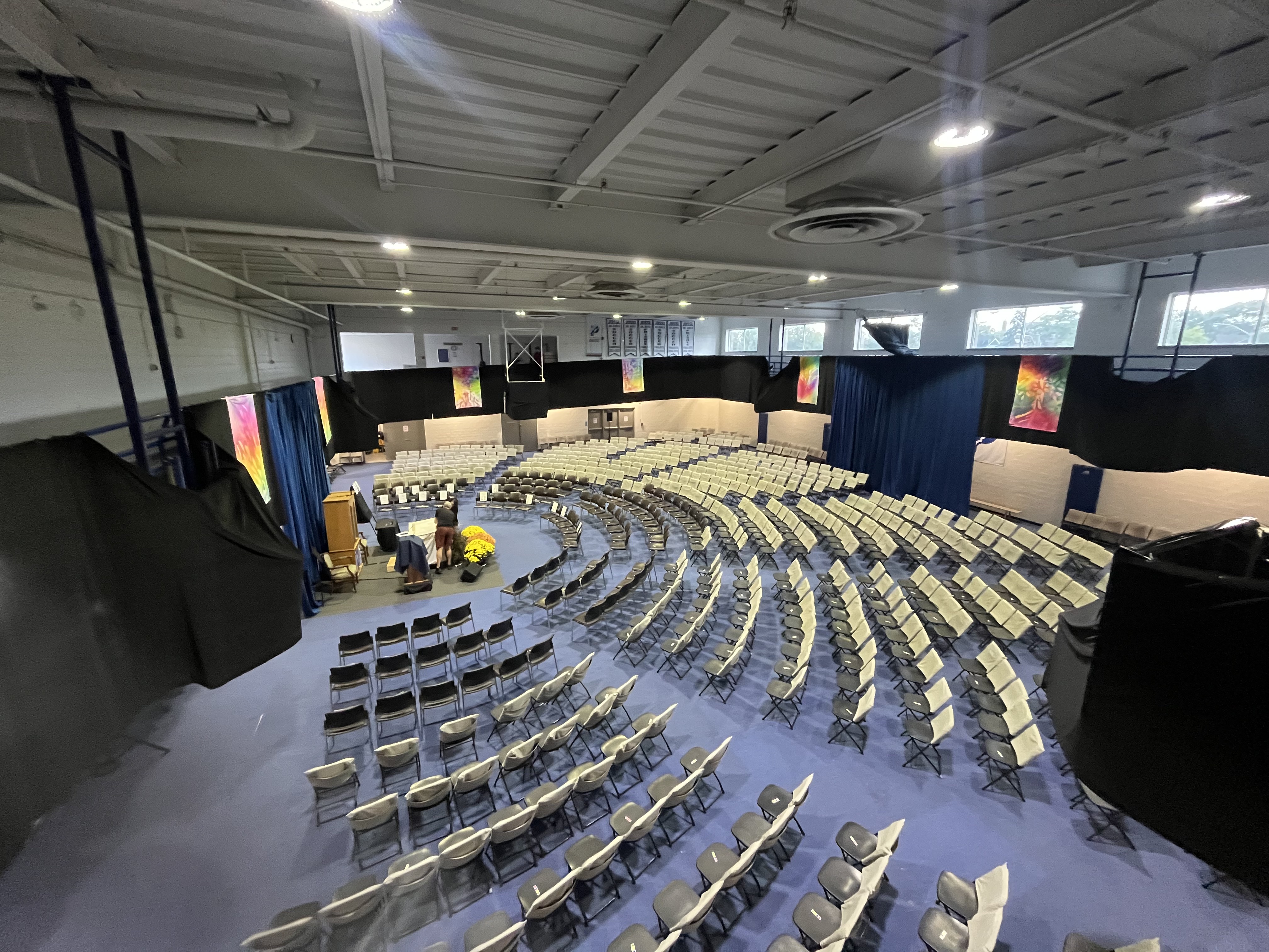 Large indoor auditorium with rows of folding chairs arranged in a semicircle facing a stage with a podium and yellow flowers.