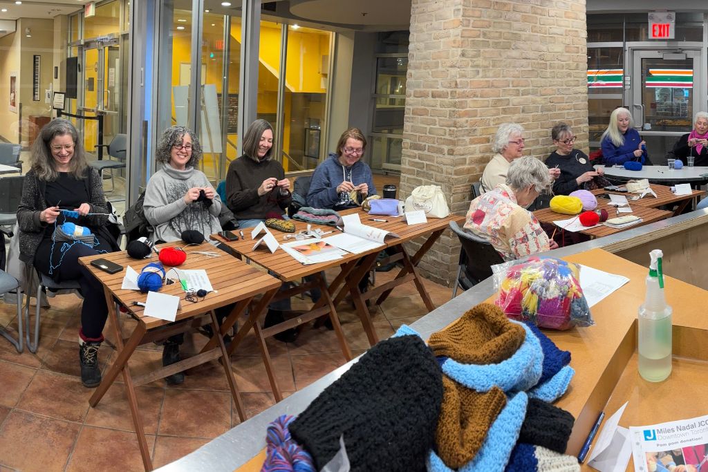 Group of women sitting in a community room, knitting with colorful yarns on wooden tables.