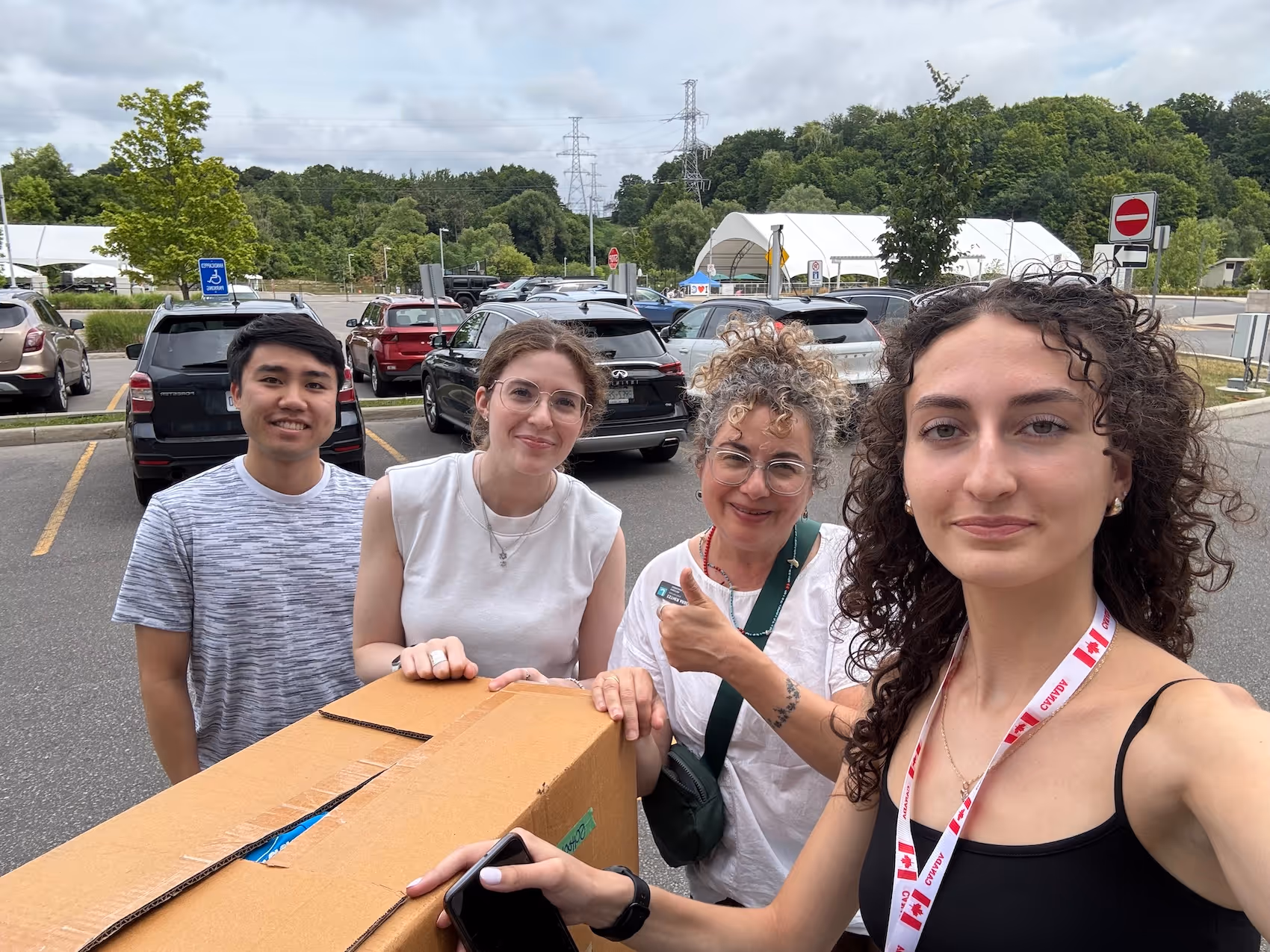 Group of four people outdoors with a large cardboard box, smiling in a parking lot with cars and trees in the background.