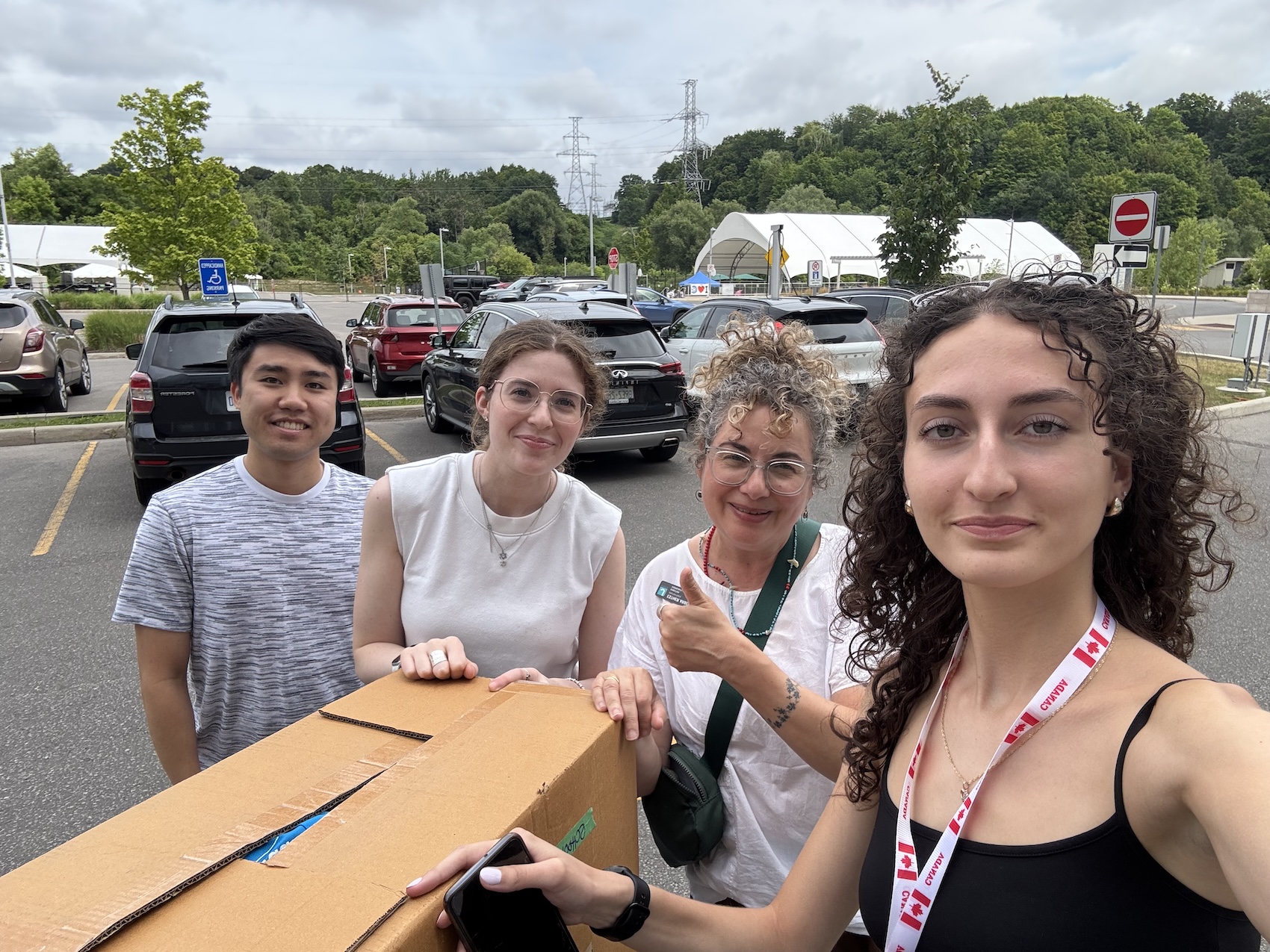 Group of four people outdoors with a large cardboard box, smiling in a parking lot with cars and trees in the background.