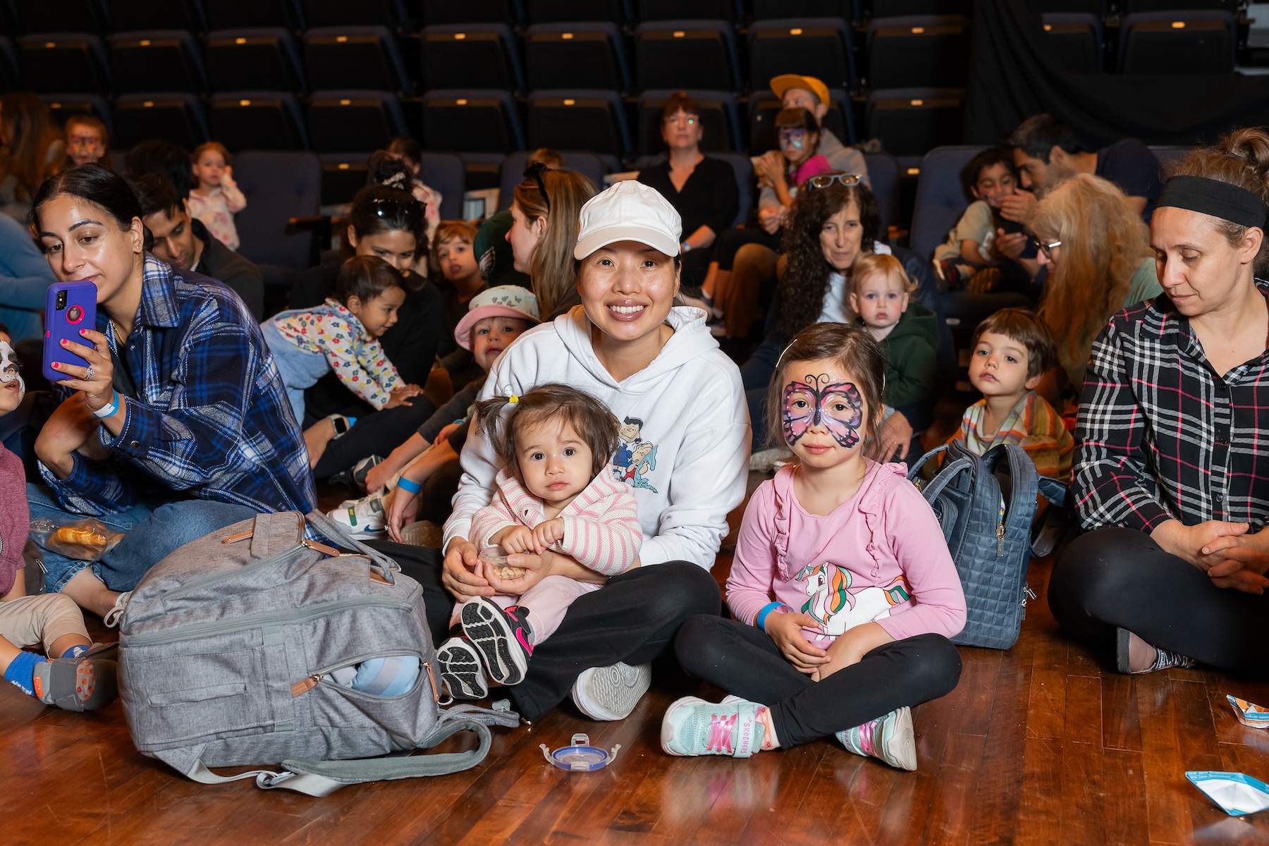 A diverse group of adults and children sitting on a wooden floor in a theater setting, with one child wearing butterfly face paint and another being held by an adult in a white cap.