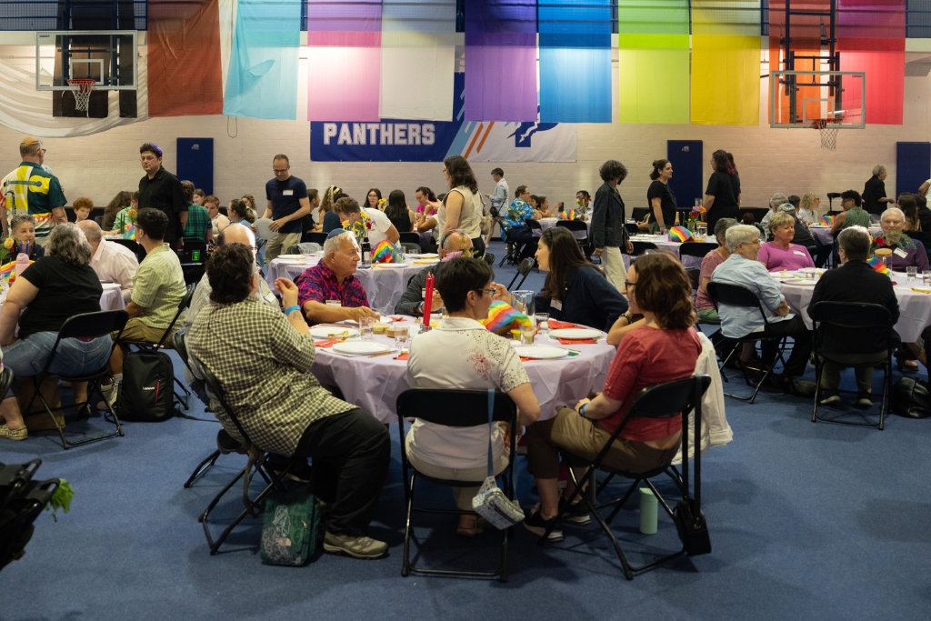 People sitting at round tables covered with light purple tablecloths in a gymnasium decorated with colorful vertical banners and a 'Panthers' banner, engaging in conversation and dining.