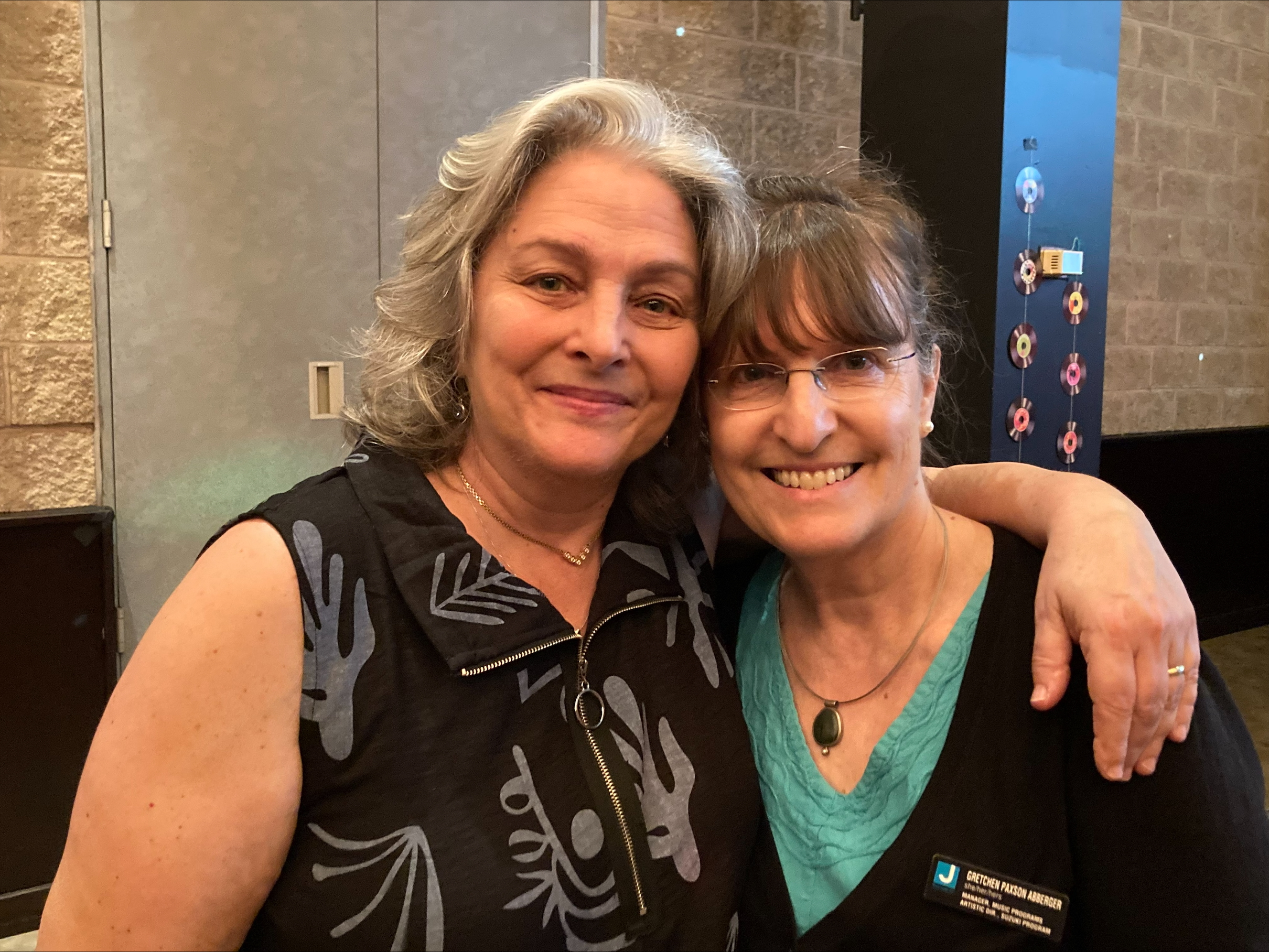 Two middle-aged women smiling, one with gray hair and patterned black top, the other wearing glasses, a blue top, and a name badge that reads Gretchen Paxson Asberger.