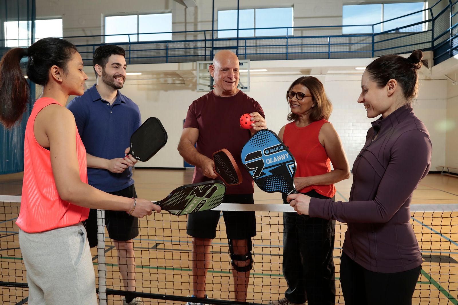 Five adults holding pickleball paddles and ball on an indoor court, smiling and preparing to play.