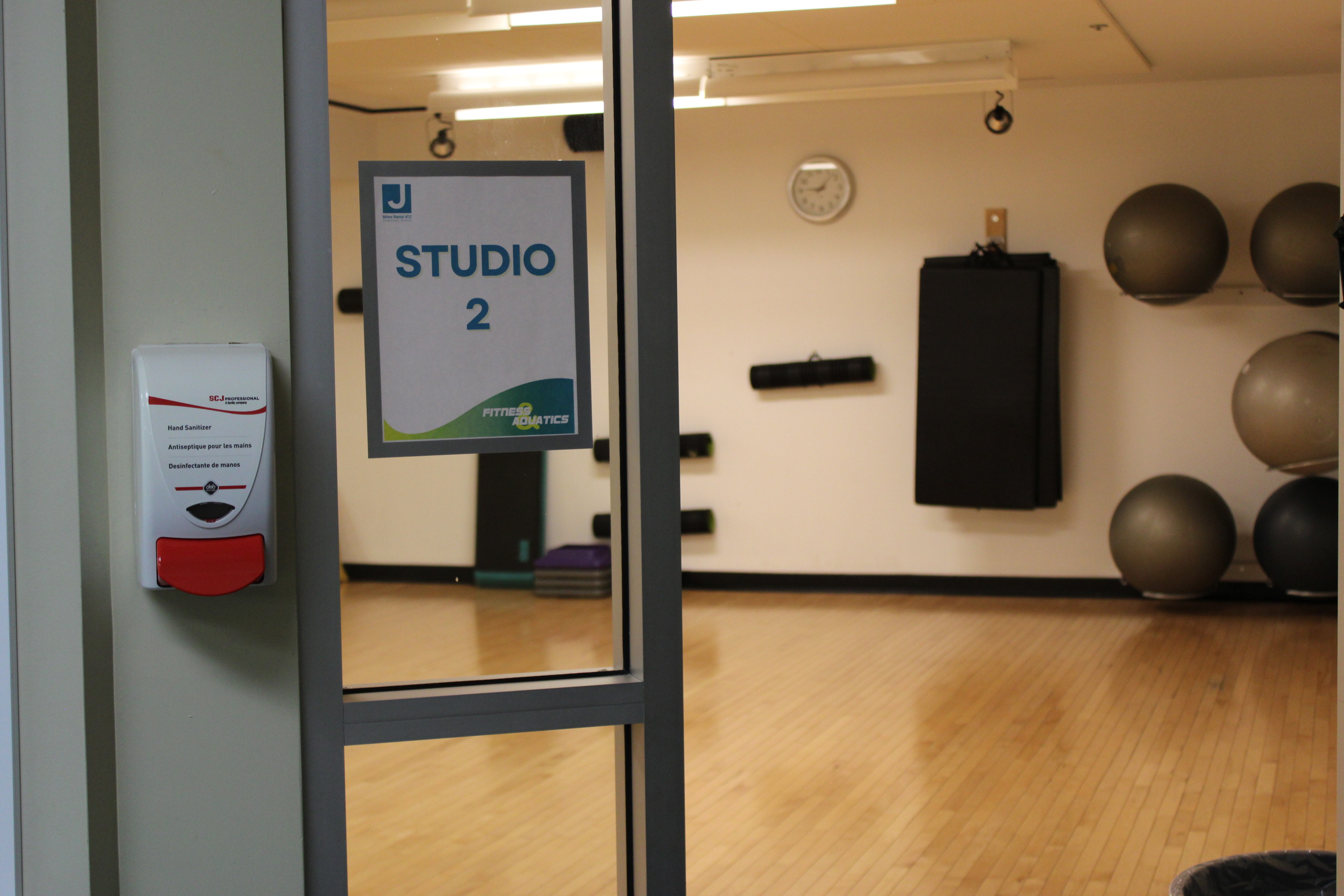 Glass door labeled Studio 2 with a hand sanitizer dispenser on the wall beside it, showing an empty indoor fitness studio with exercise balls, mats, and a clock on the wall.