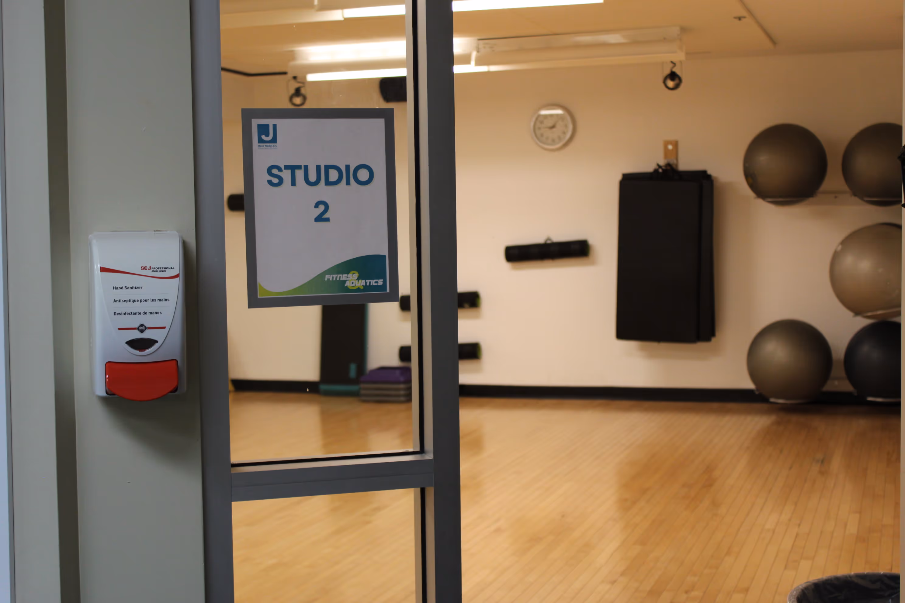 Glass door labeled Studio 2 with a hand sanitizer dispenser on the wall beside it, showing an empty indoor fitness studio with exercise balls, mats, and a clock on the wall.
