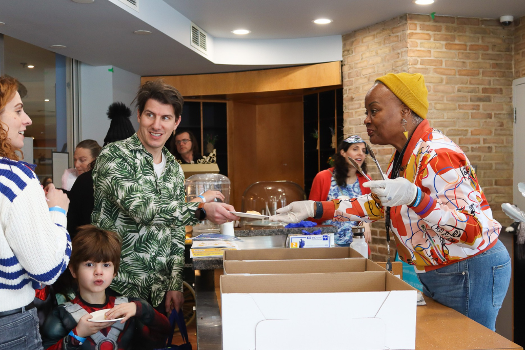 Volunteer wearing gloves and a colorful jacket serving food on a plate to a man with a green leaf-patterned shirt at a counter.