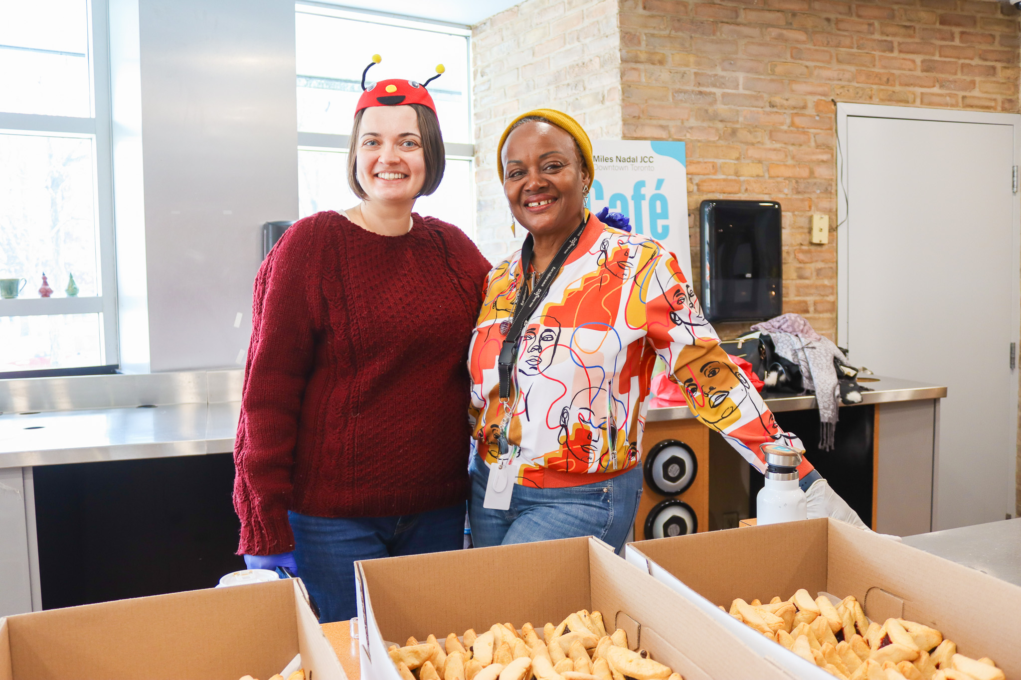 Two women smiling in a kitchen-like setting with large boxes of pastries in front of them; one woman wears a ladybug headband and red sweater, the other a colorful shirt and yellow beanie.