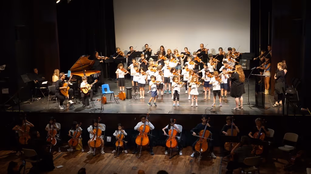 Children and adults performing a music concert with violins, cellos, guitars, and a piano on stage.