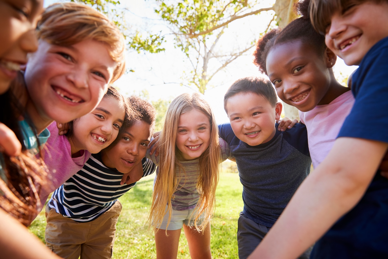 Group of diverse children outdoors in a circle with arms around each other, smiling at the camera.