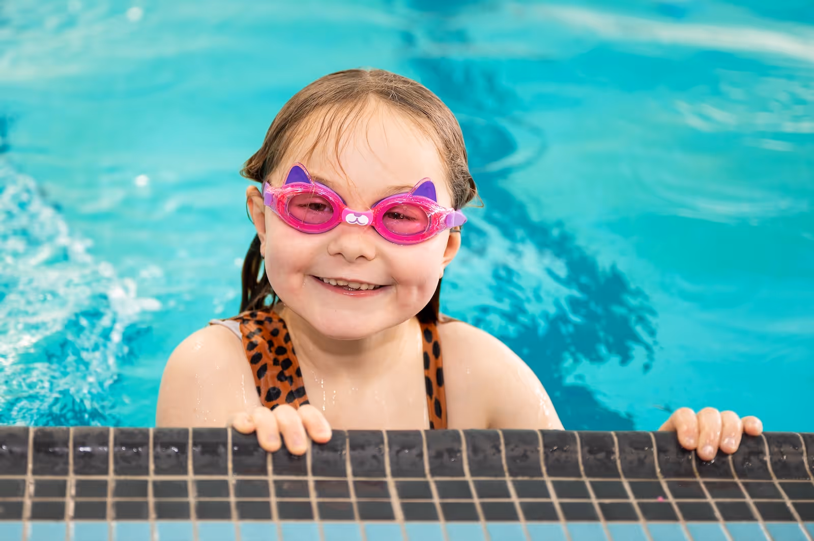 Smiling young girl wearing pink cat-shaped swimming goggles holding onto the edge of a pool.