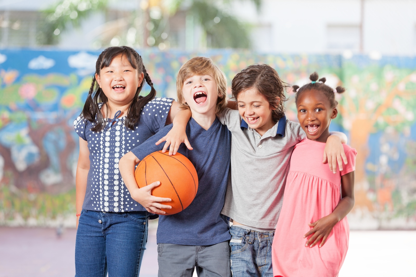 Group of four diverse children smiling and laughing, standing with arms around each other, one holding a basketball.