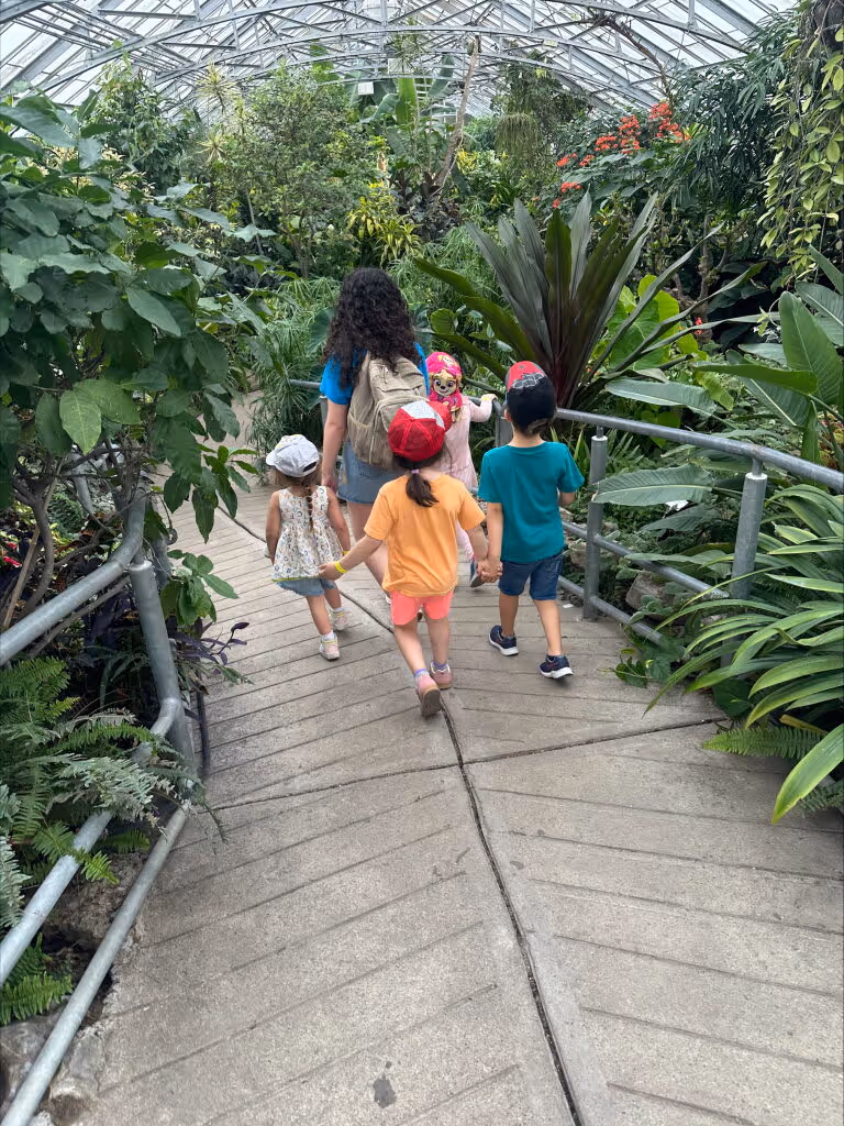 Four children walking hand-in-hand along a path in a lush greenhouse filled with green plants and flowers.
