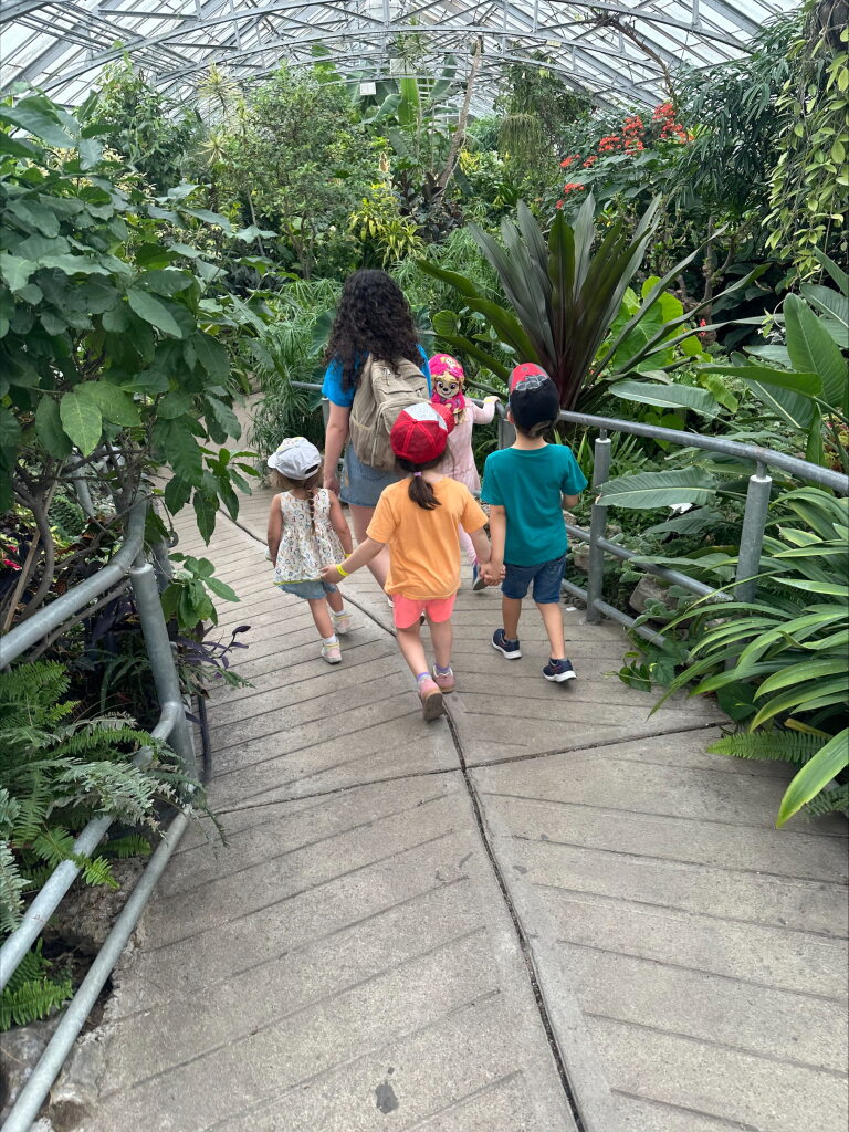 Four children walking hand-in-hand along a path in a lush greenhouse filled with green plants and flowers.