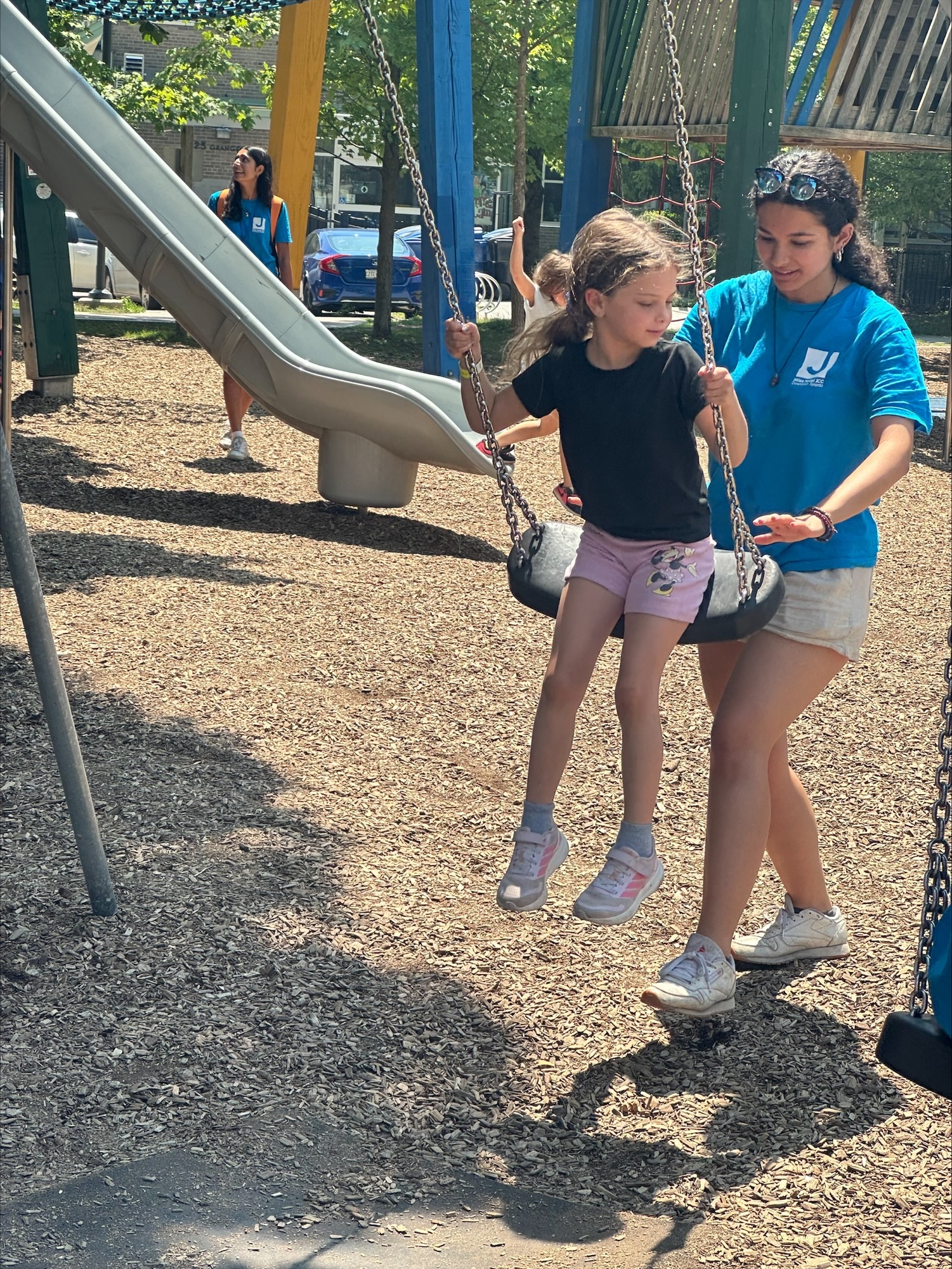 A woman in a blue shirt pushes a girl in pink shorts on a swing at a playground on a sunny day.