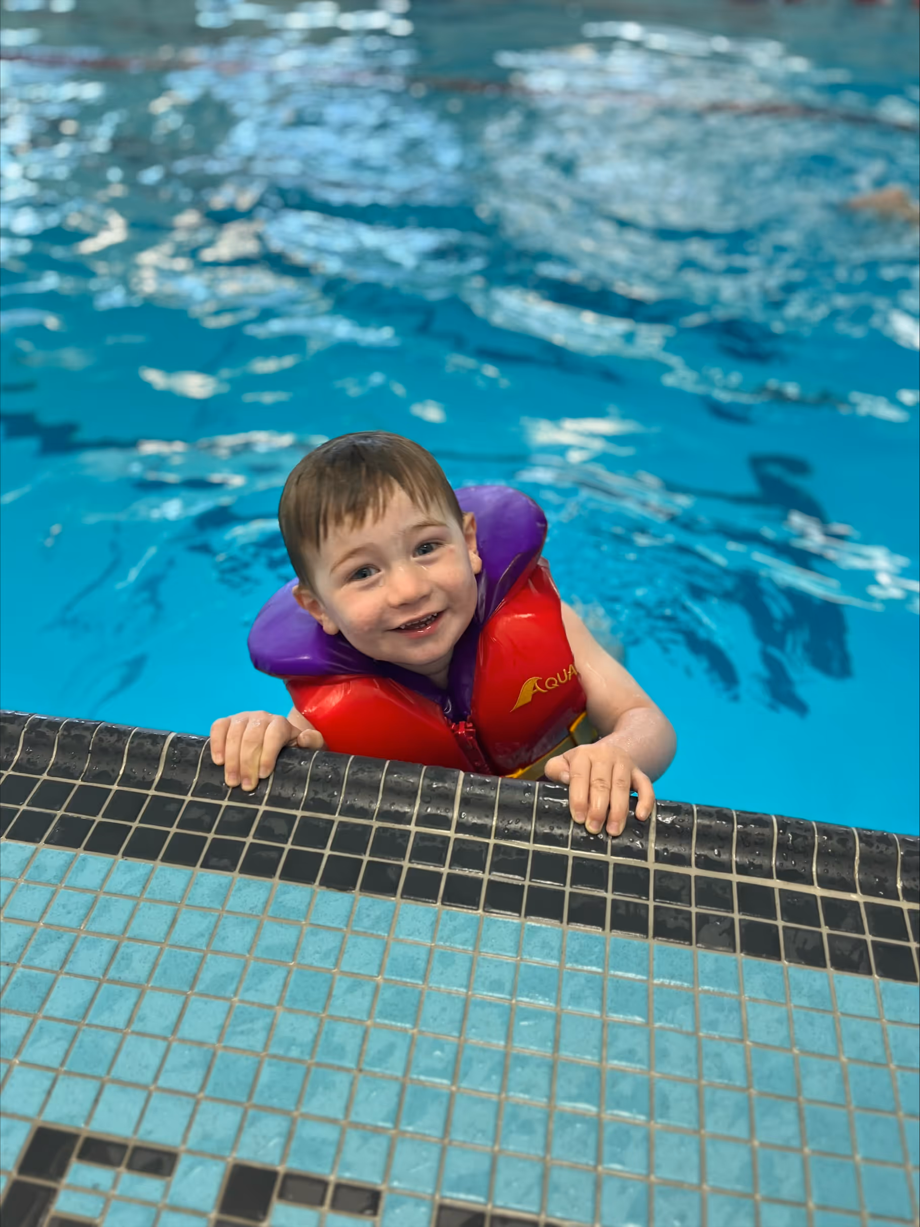 Young boy smiling while holding onto the edge of a swimming pool wearing a red and purple life jacket.