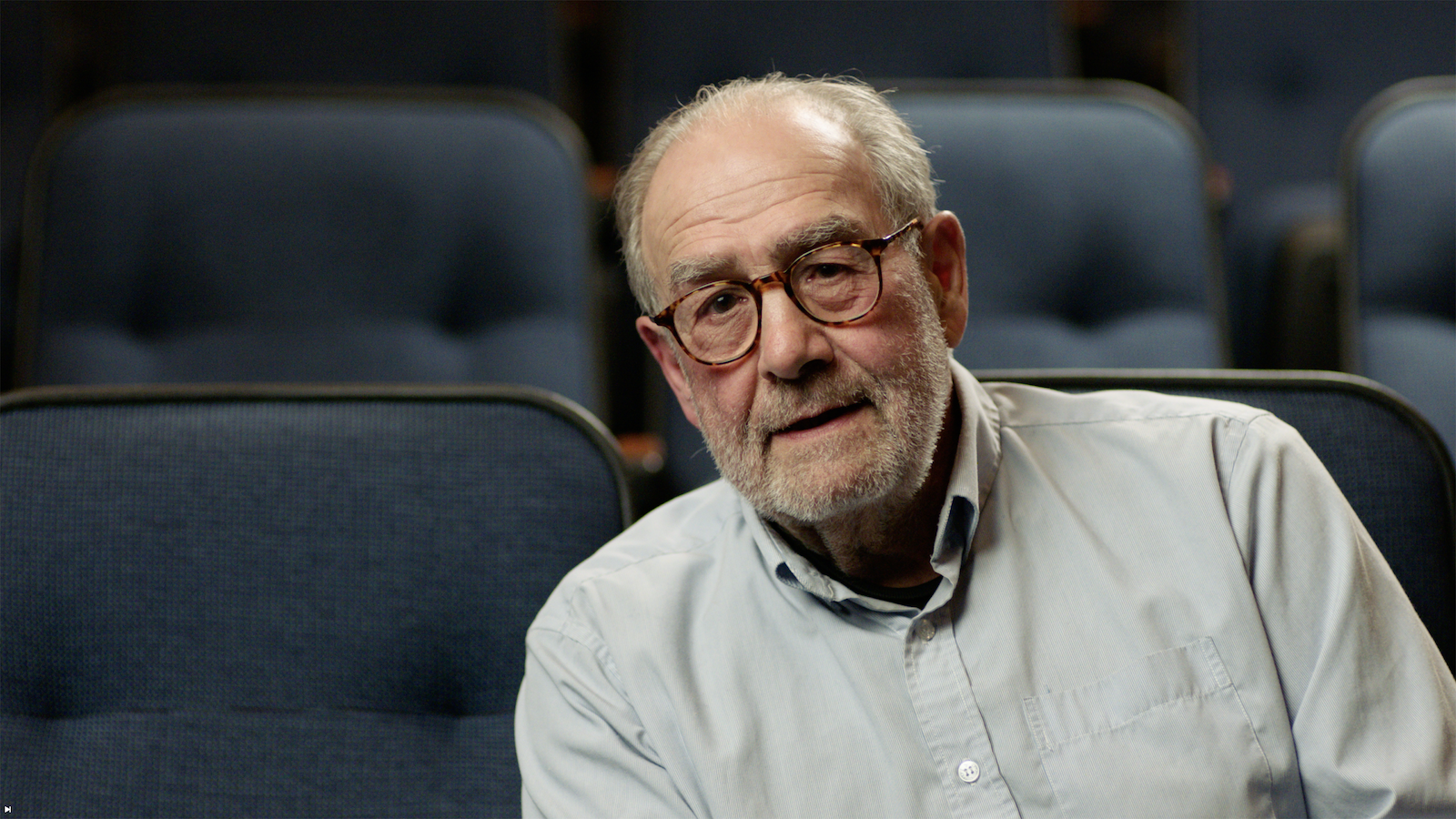 Elderly man with glasses and a light blue shirt sitting in a theater or auditorium with empty blue seats around him.