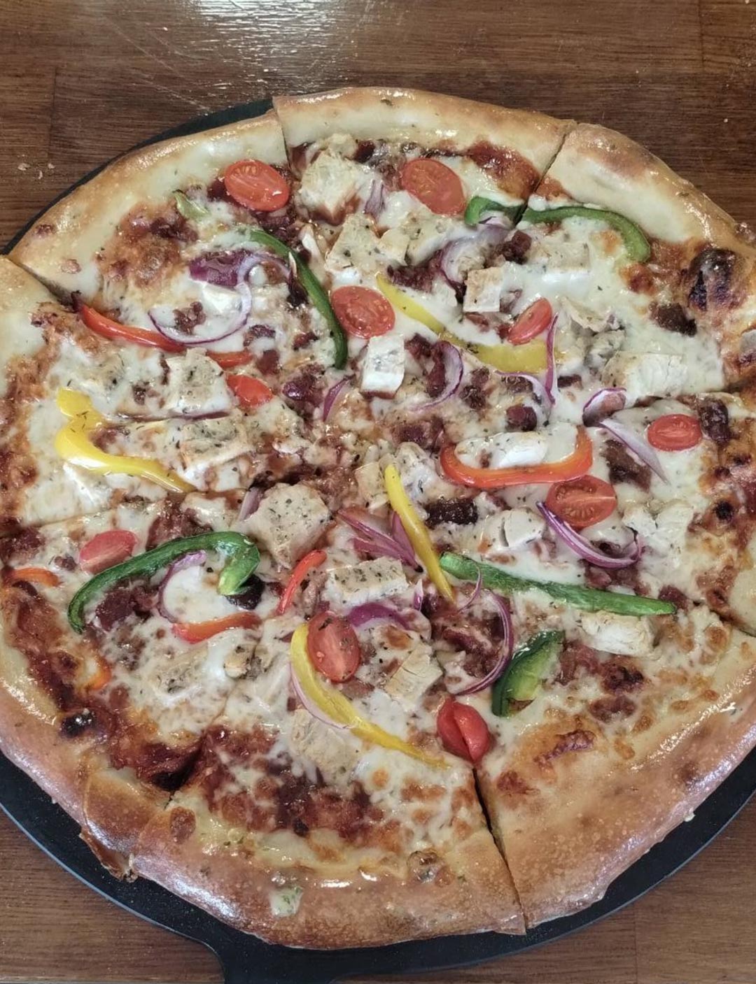 A freshly baked pizza with a golden crust, topped with sliced bell peppers, red onions, cherry tomatoes, and chunks of chicken on a wooden table.