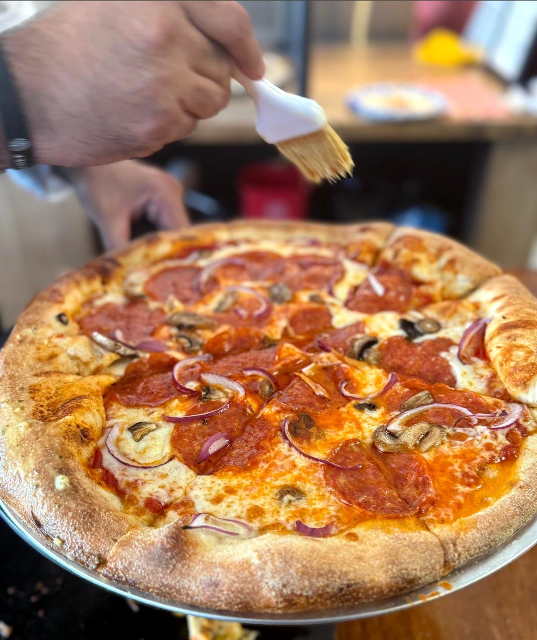 Close-up of a freshly baked pizza topped with pepperoni, red onions, and mushrooms. A hand is brushing additional oil or sauce on top, suggesting a final touch of preparation.