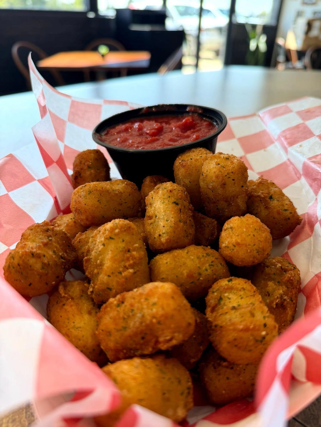 Basket of crispy tater tots served on red and white checkered paper with a small black cup of salsa for dipping, in a casual dining setting.