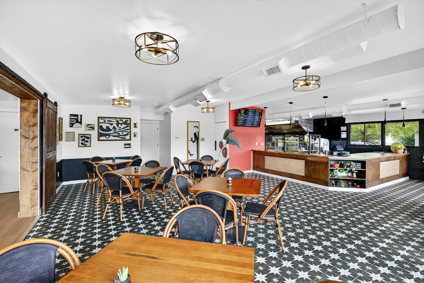 Bright, modern café interior with geometric black-and-white tiled floor, wooden tables, and wicker chairs. Art decorates the walls, and a menu board hangs near a counter.