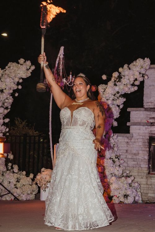 Bride in a white lace wedding dress holding a flaming torch and smiling at an outdoor nighttime celebration with floral decorations.
