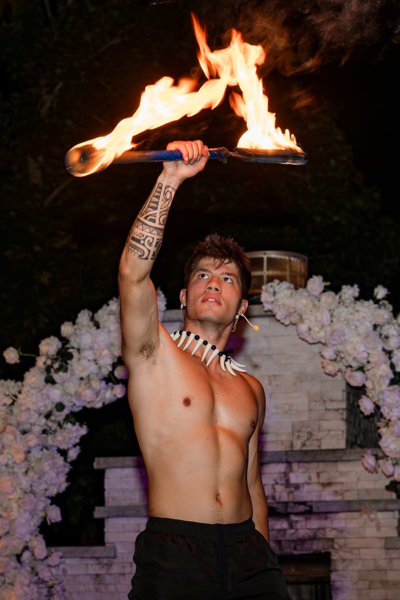 Shirtless man with tribal arm tattoo and necklace holding a flaming torch above his head, surrounded by white flowers.