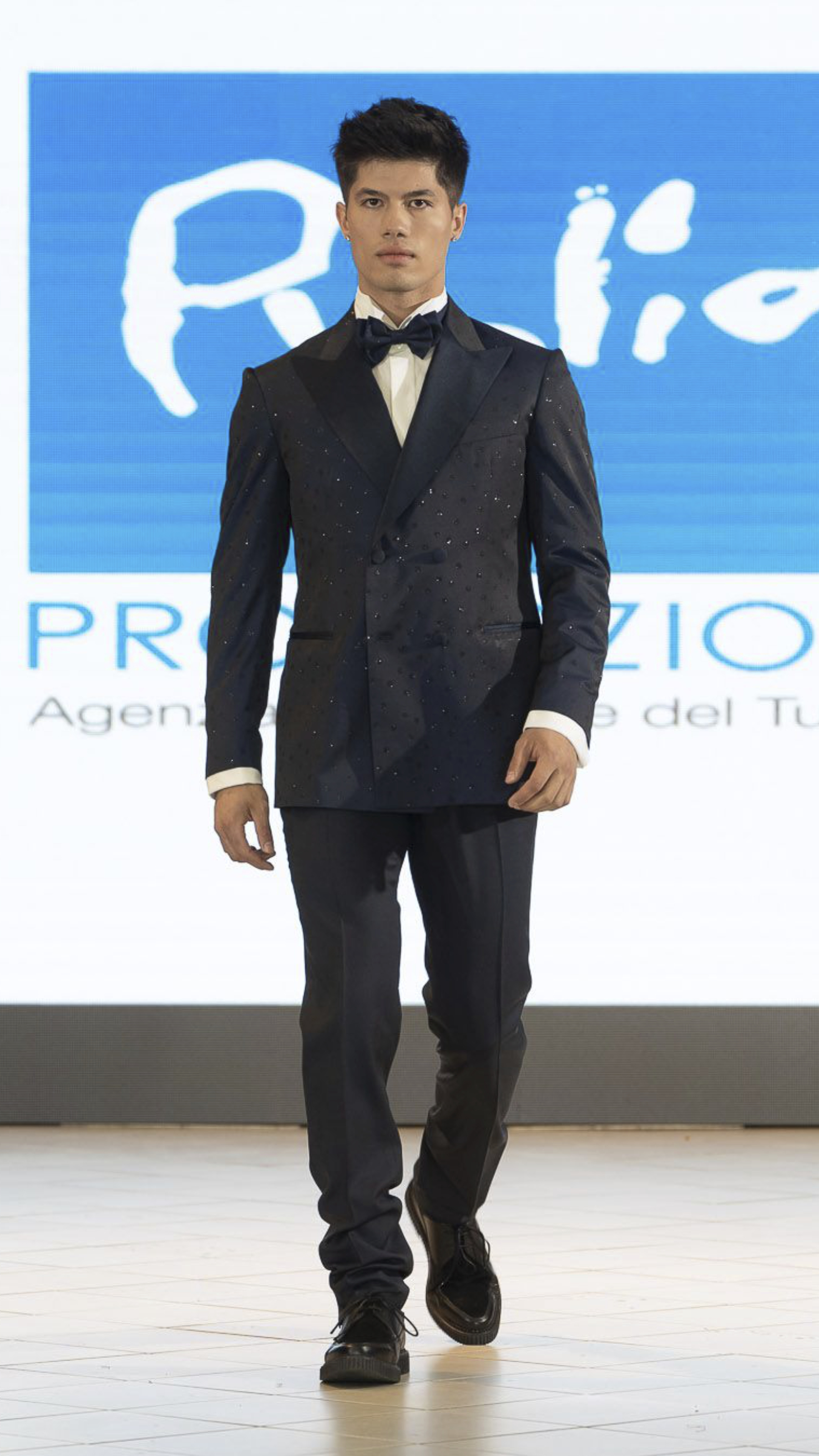 Male model walking on runway wearing a dark suit with a bow tie and black shoes.