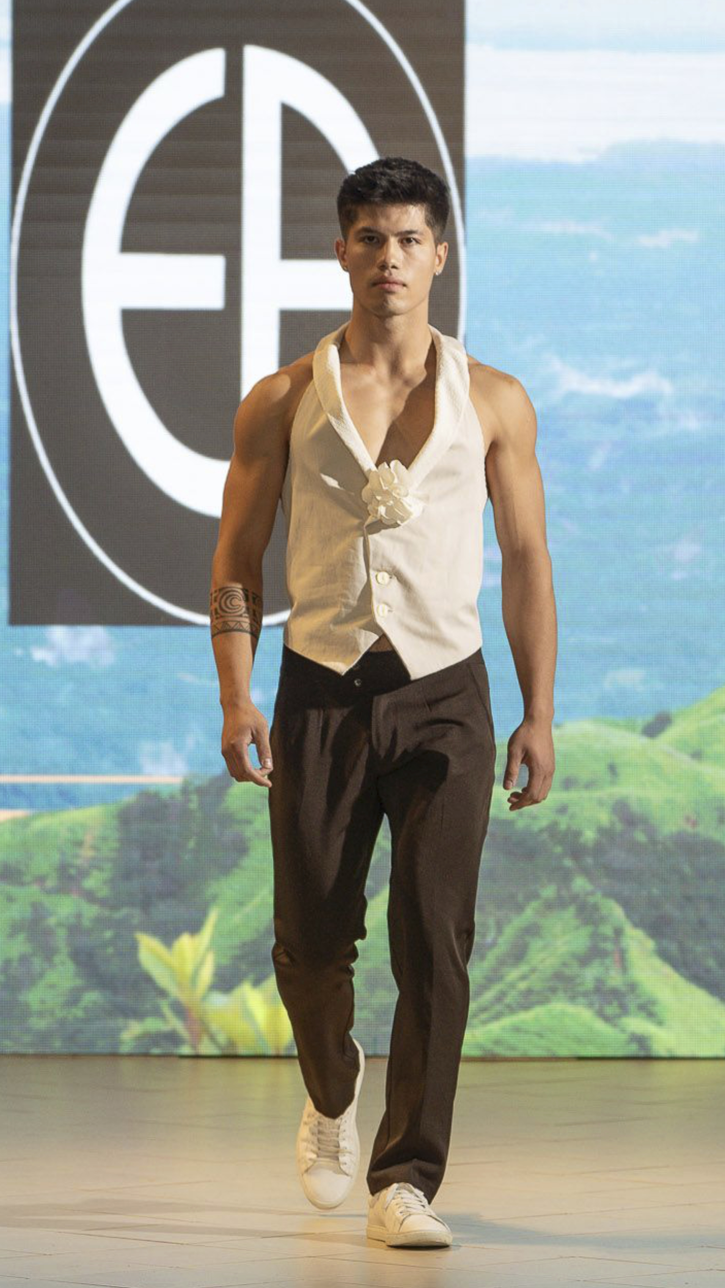 Male model walking on runway wearing a sleeveless white vest with a flower detail, black pants, white sneakers, and an arm tattoo, against a backdrop of green hills and blue sky.
