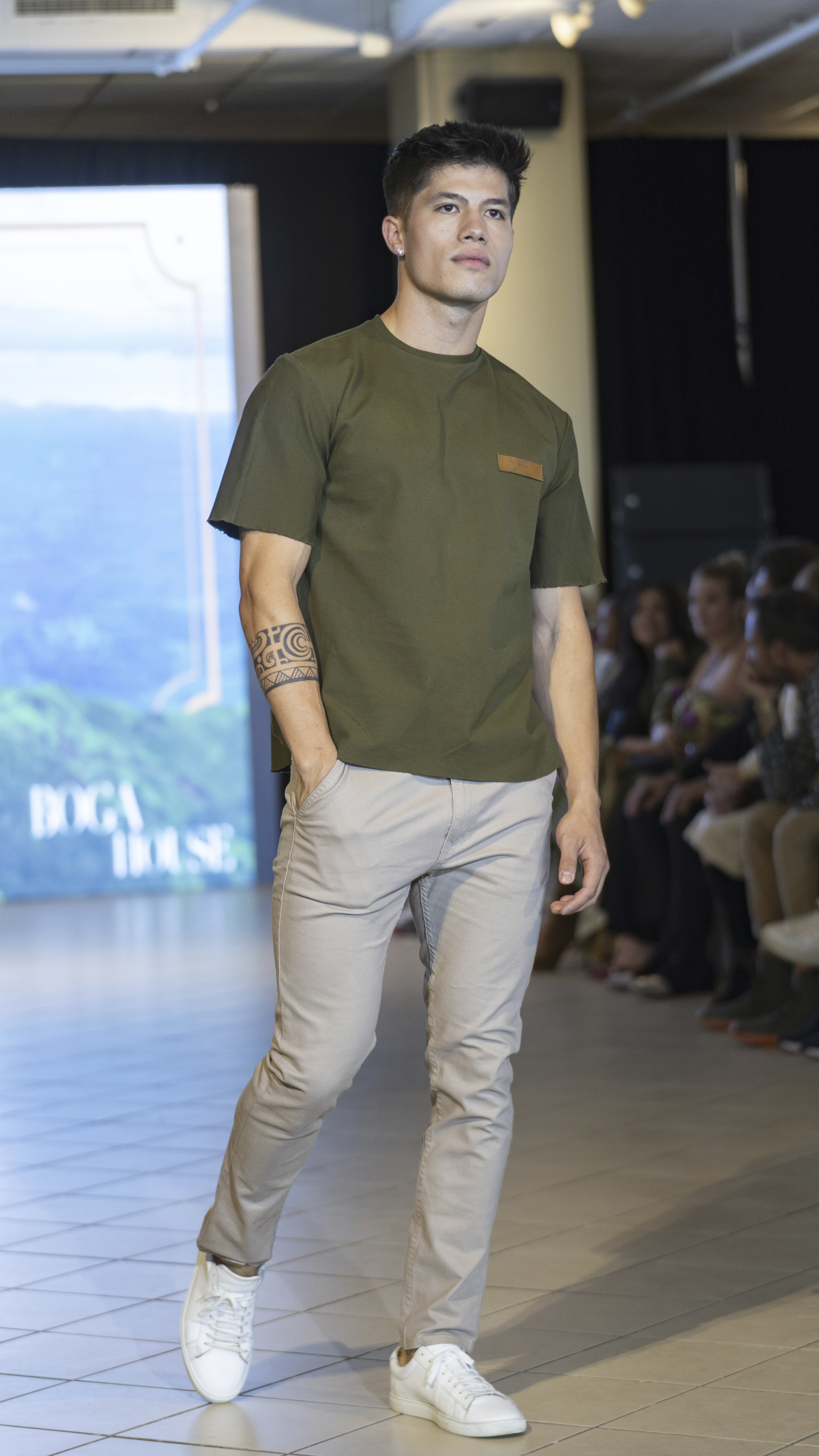 Male model with tattoo on right forearm walking in olive green t-shirt, beige pants, and white sneakers on fashion runway.